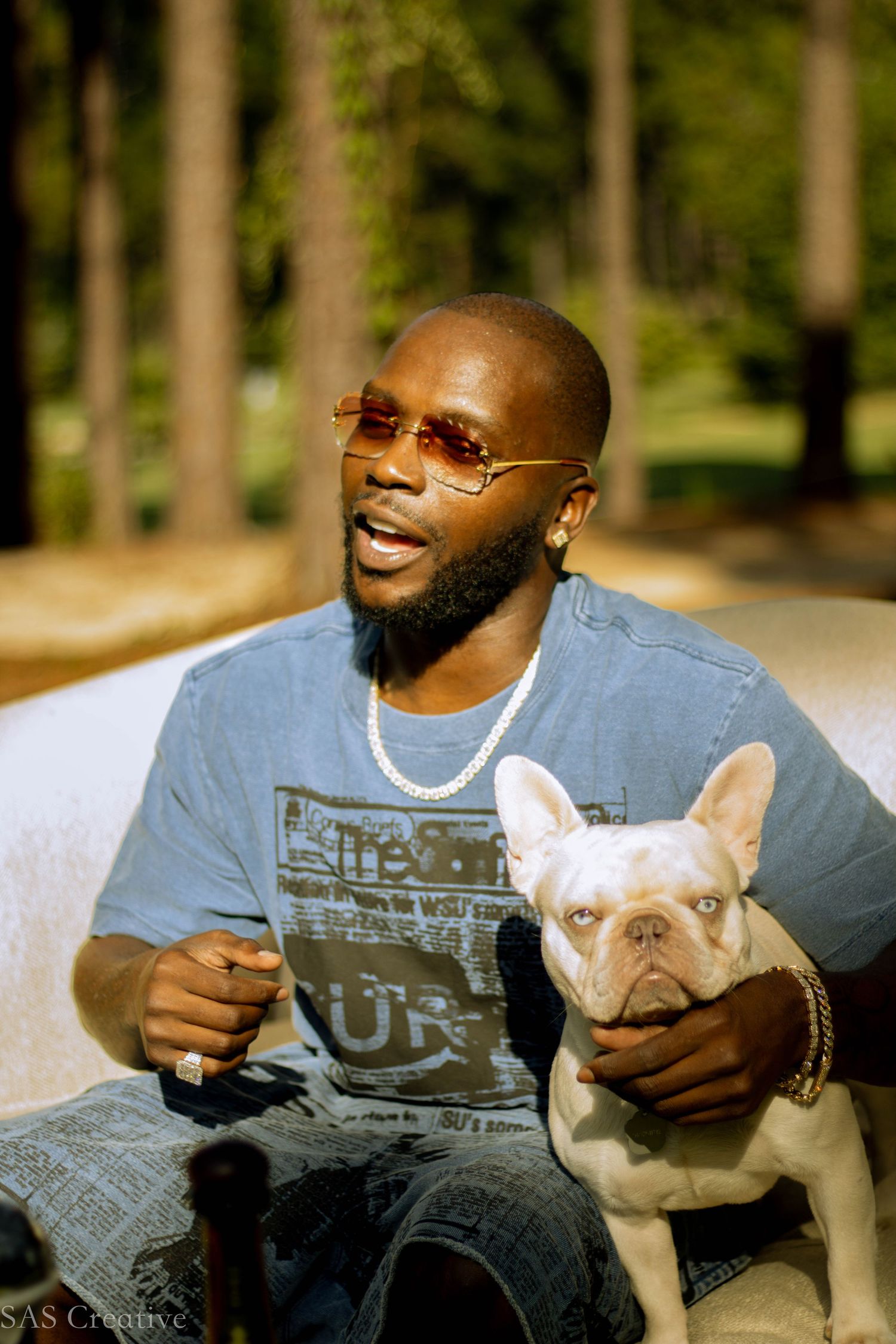 Bearded man wearing sunglasses and white chain necklace smiles while holding a white French Bulldog outdoors.