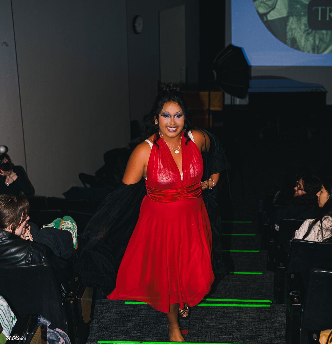 A woman in a red halter midi dress walks down an auditorium aisle with bold eye makeup.