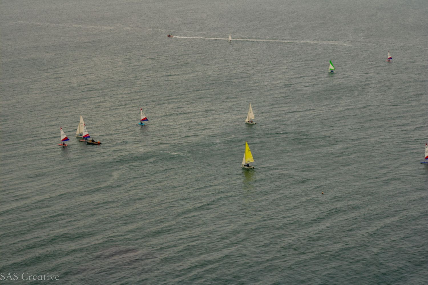 Aerial view of colorful sailboats with bright sails racing across calm open water.