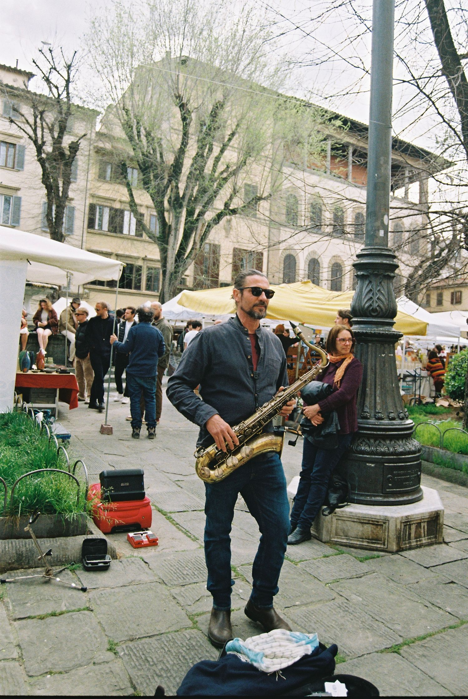A street musician plays saxophone at an outdoor market in a European city square lined with white vendor tents.