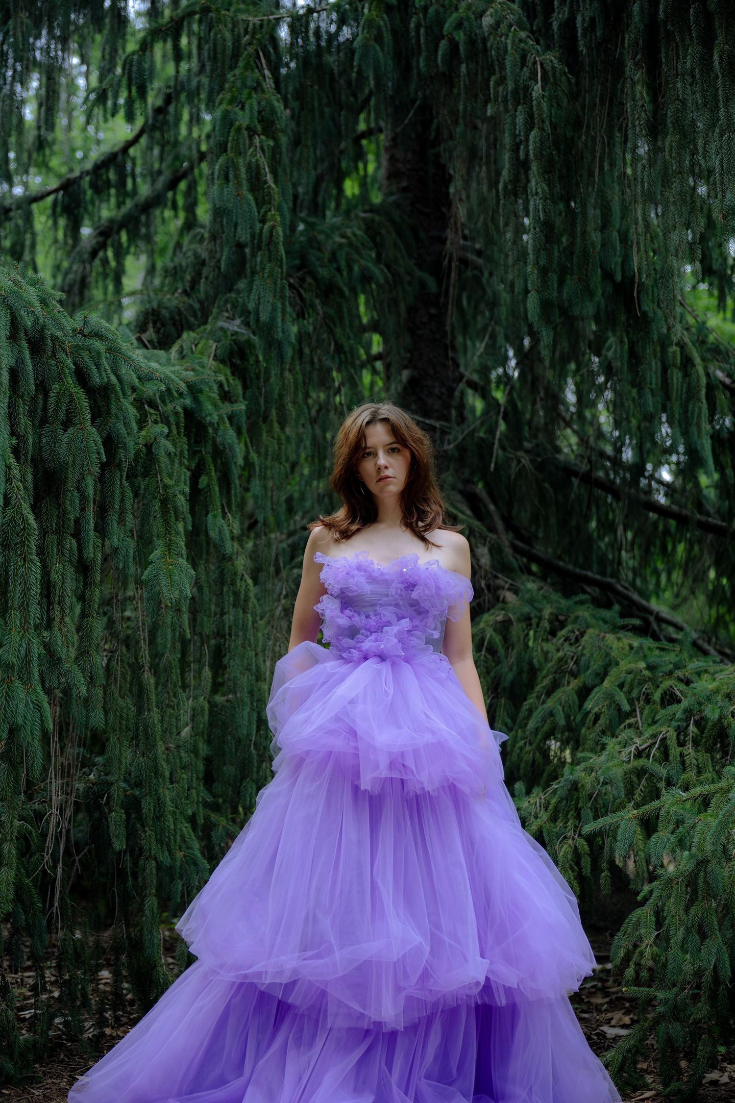 Woman in lavender tulle ball gown poses among lush green weeping pine trees in an enchanting forest setting.