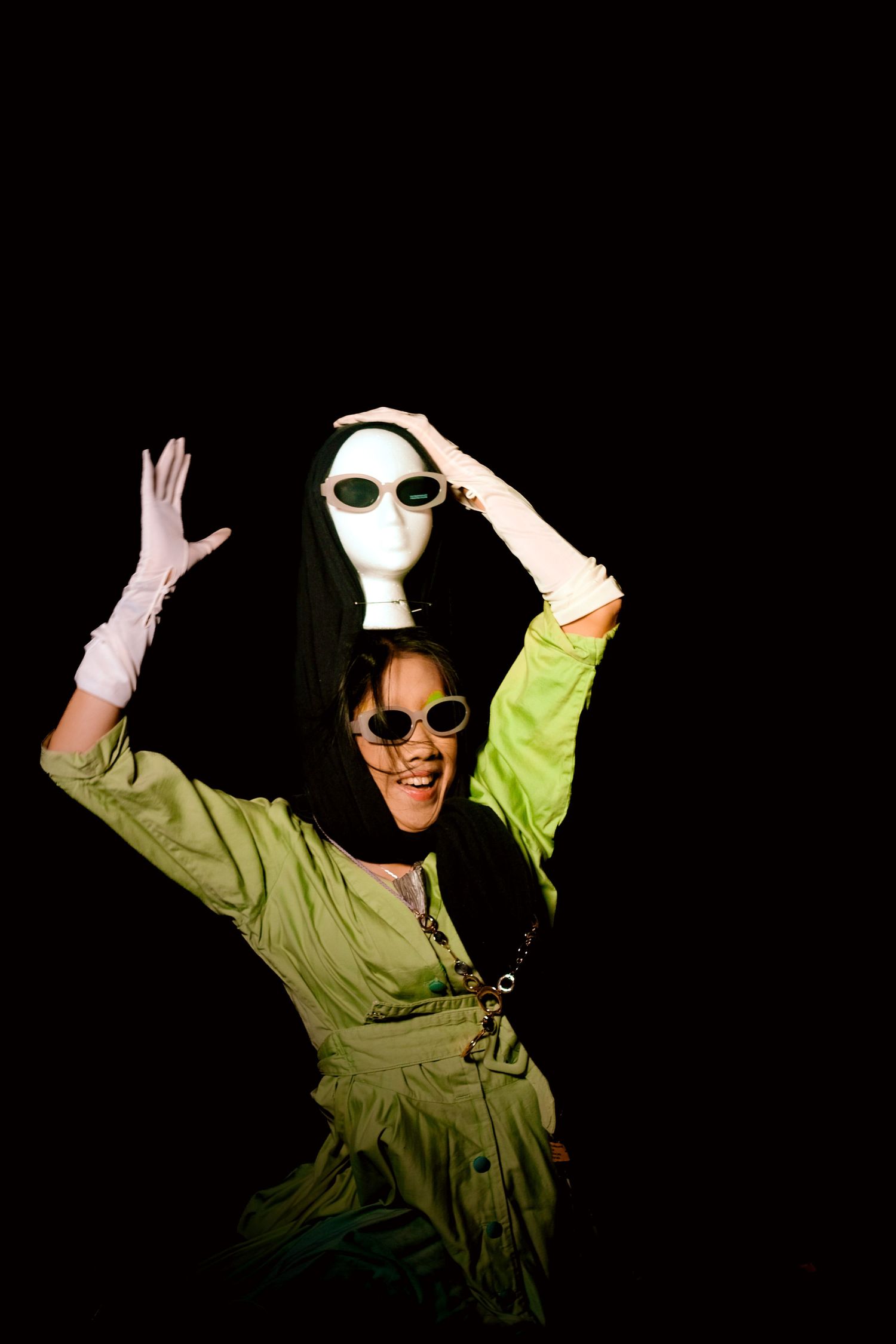 Woman in green dress holding a white mannequin head above her on a black background.