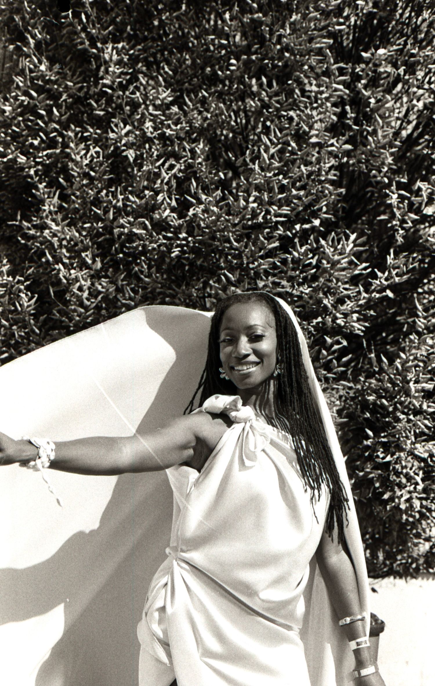 Black and white photo of a smiling Black woman in a white draped outfit standing near lush bushes.