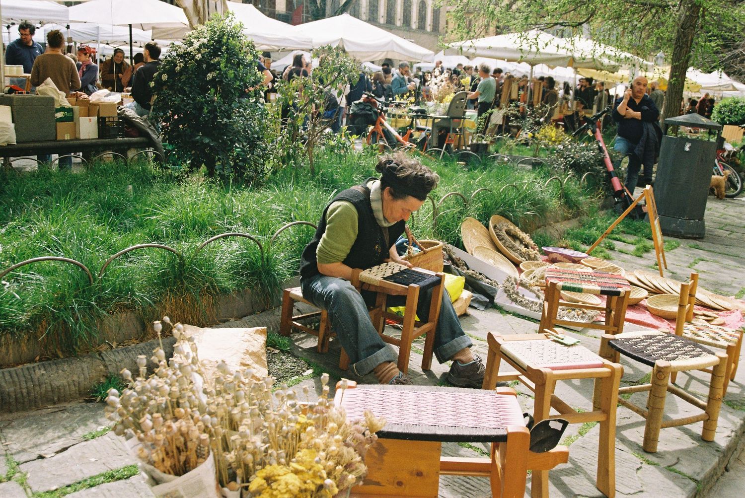 Craftsman weaving a chair seat at an outdoor artisan market surrounded by wooden furniture.