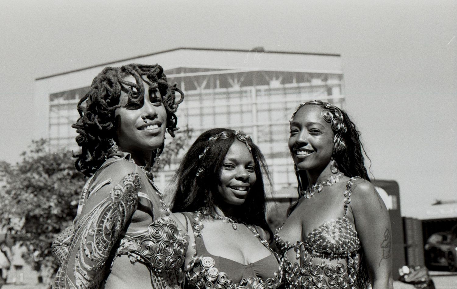 Black and white photo of three smiling young Black women in decorative outfits at an outdoor event.