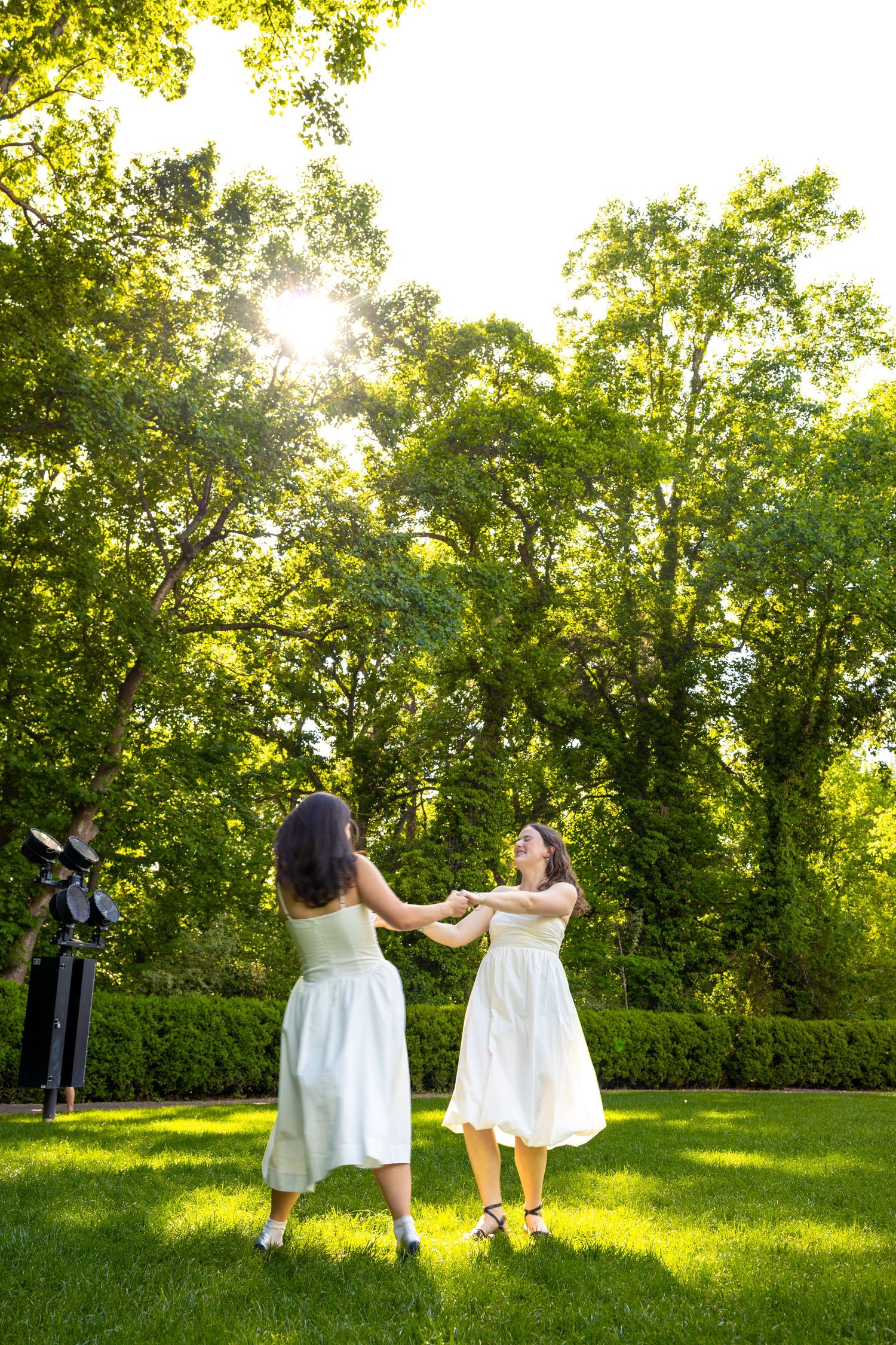 Two women in white dresses hold hands and spin joyfully on a sunny green lawn surrounded by lush trees.