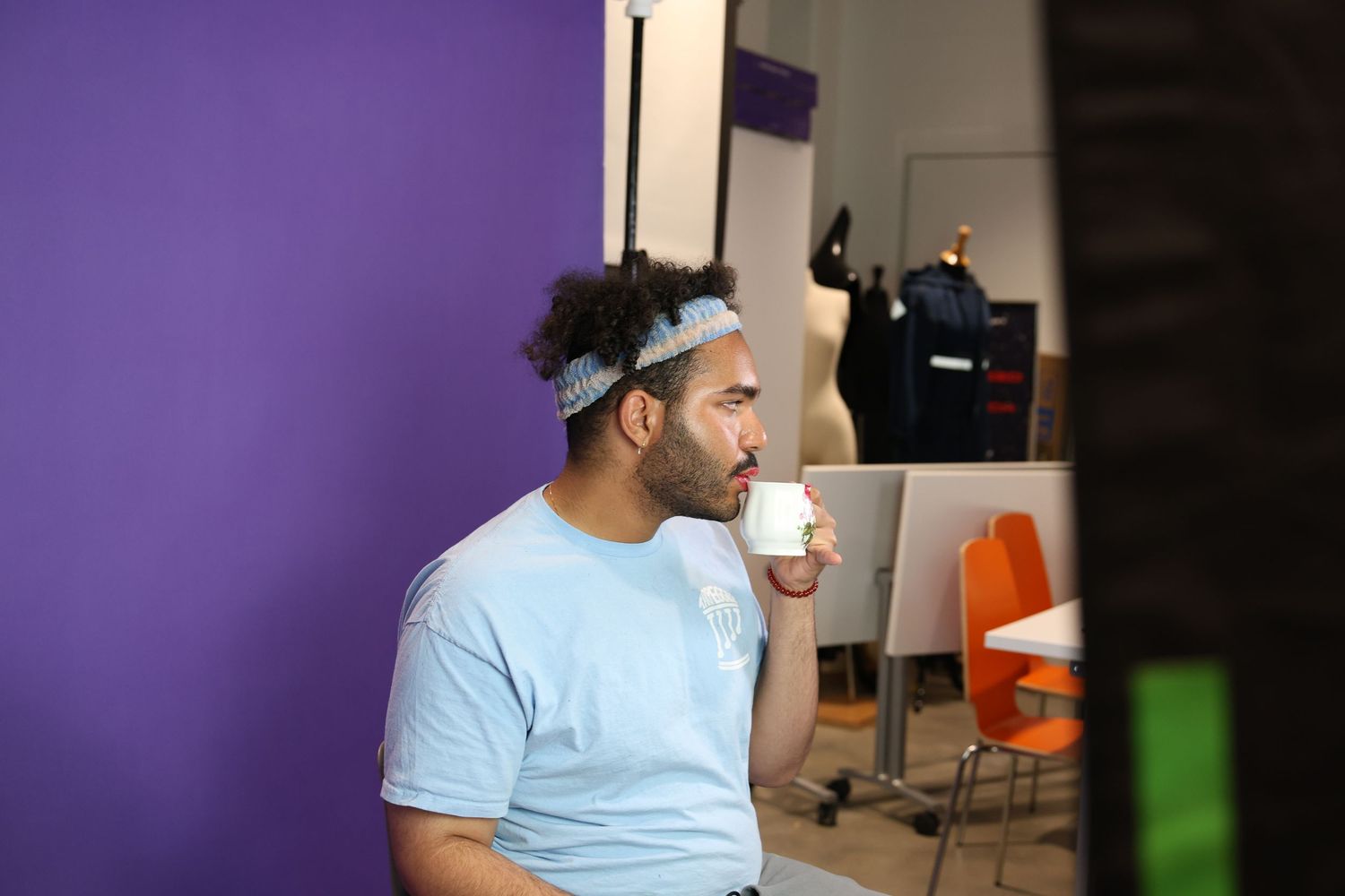 Man in light blue t-shirt drinks coffee while sitting in front of a purple wall in a studio.