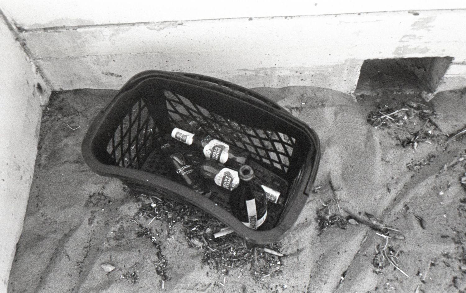 Black and white photo of bottles in a plastic crate sitting on sand against a concrete wall.