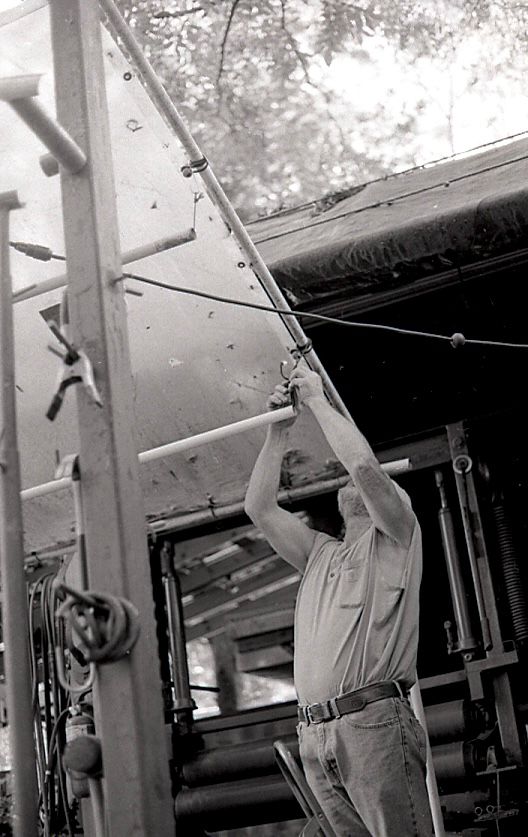 Black and white photo of a man reaching up to adjust a metal structure outdoors.