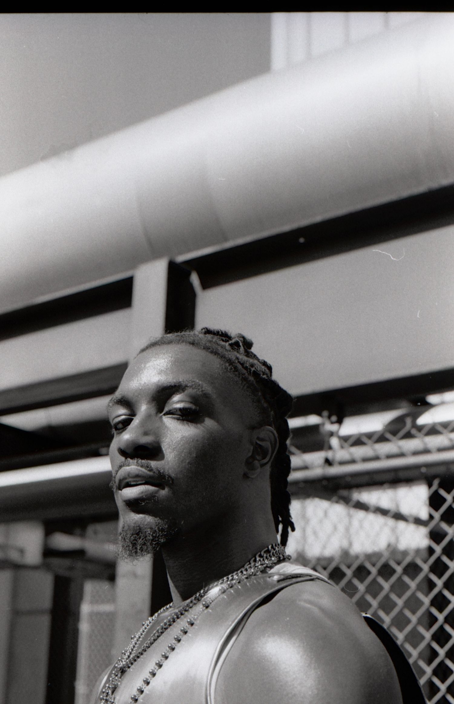 Black and white portrait of a Black man with braided hair and chain necklaces in an urban setting.