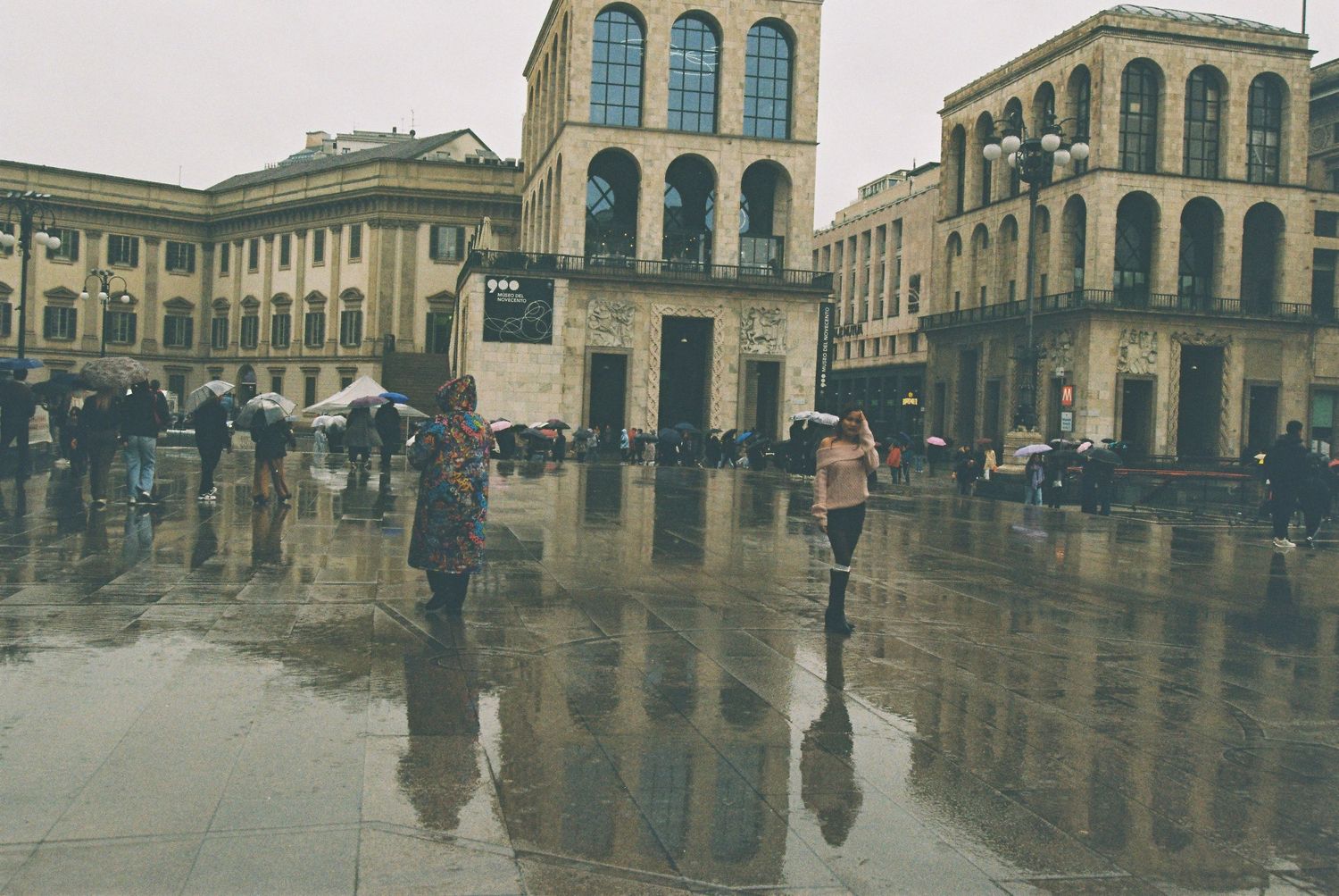 Tourists with umbrellas cross a rain-soaked piazza reflecting neoclassical buildings on an overcast day.