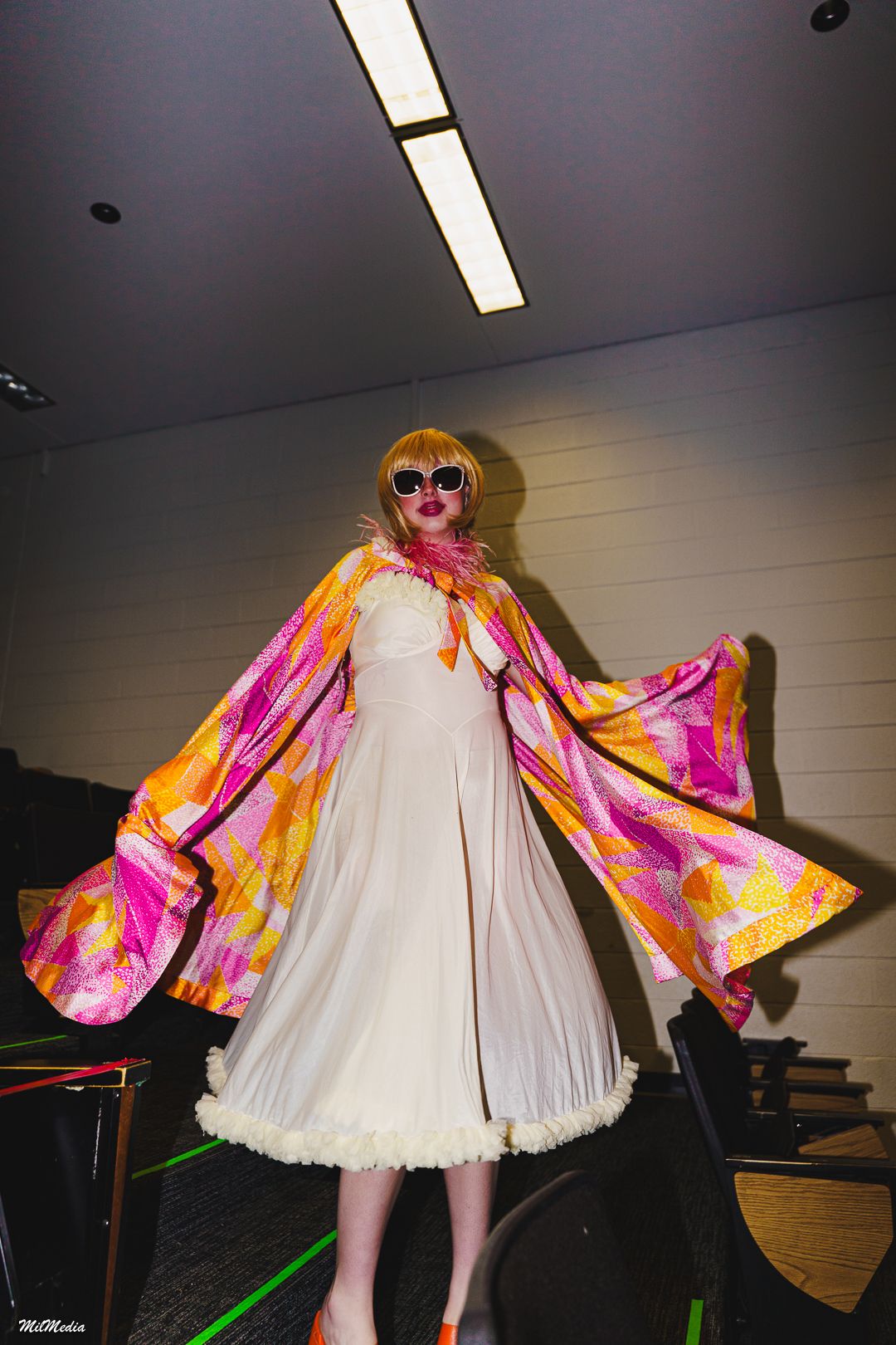 Model poses in white dress with dramatic pink and orange printed cape, blonde wig, and sunglasses at fashion show.