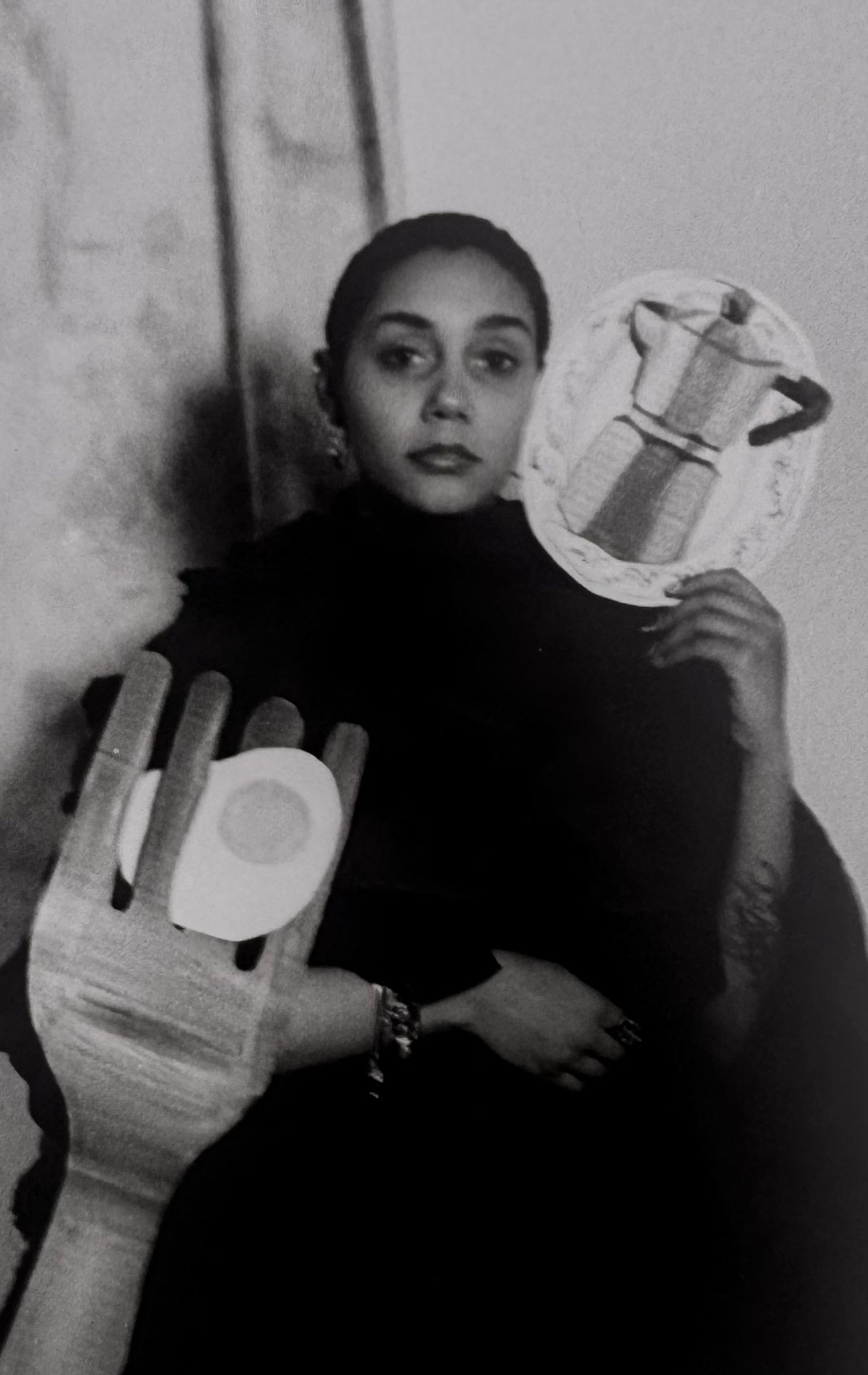 Black and white photo of a woman in a turtleneck holding artistic cutouts of a hand and moka pot.