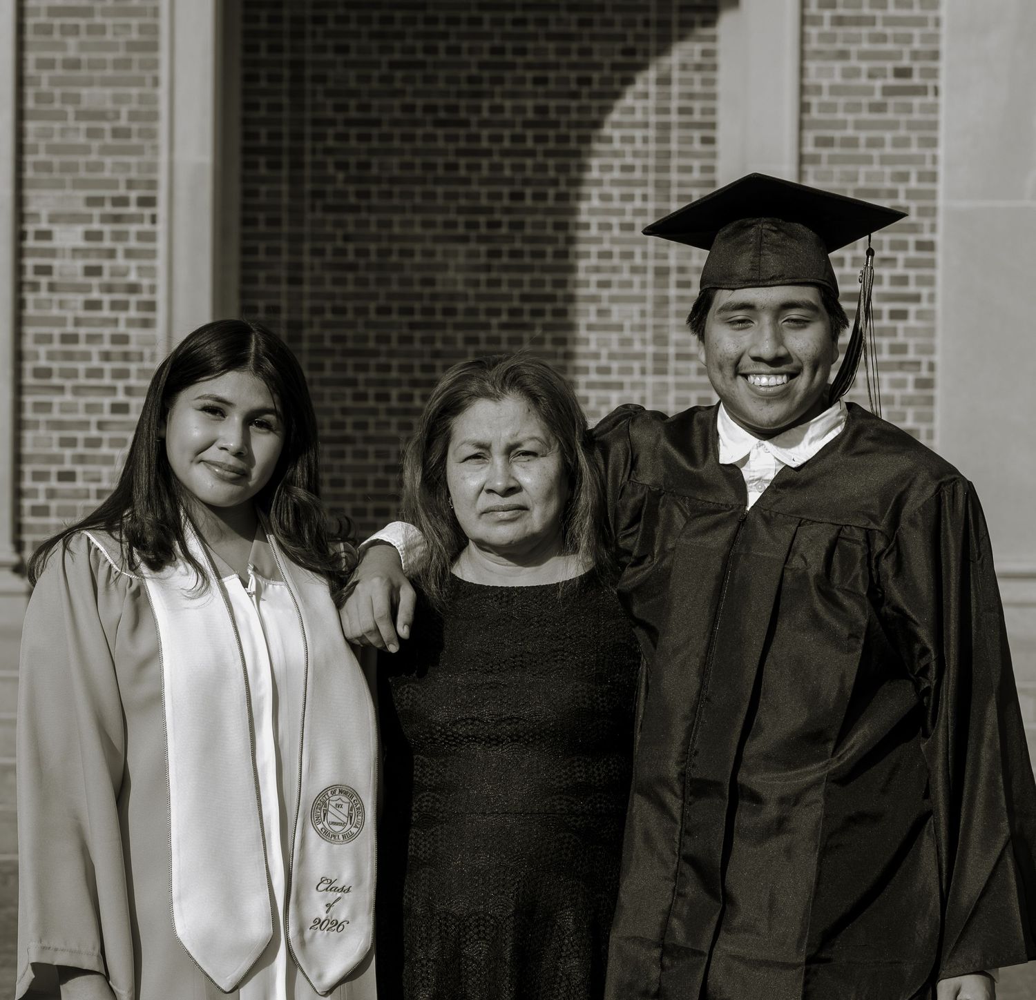 Black and white photo of two graduates in cap and gown posing with a woman in front of brick building.