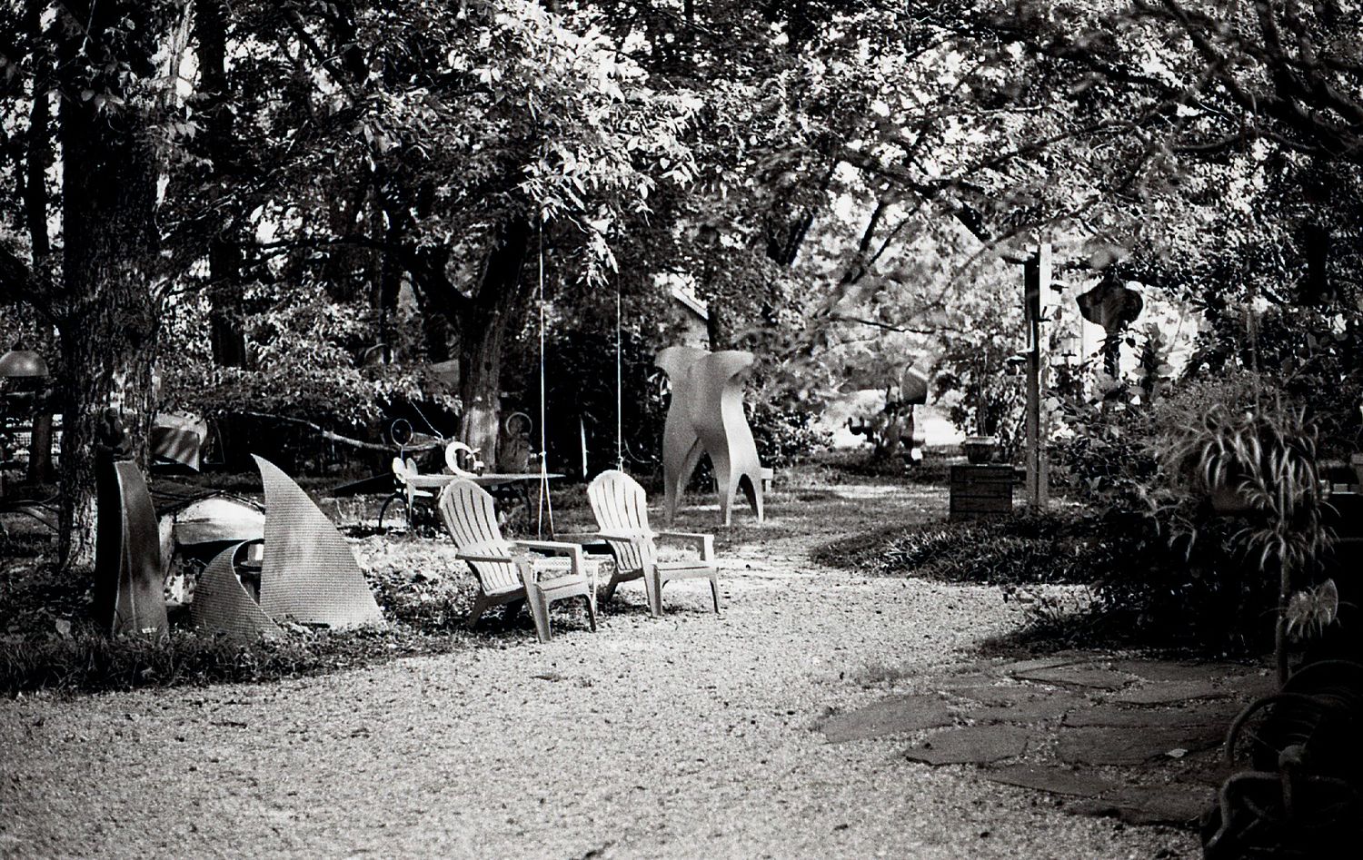 Black and white garden scene with Adirondack chairs and abstract metal sculptures among trees.
