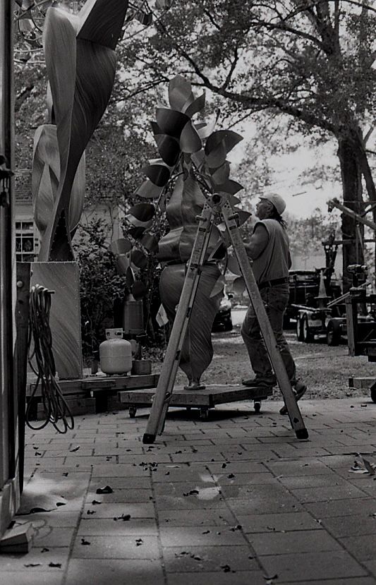 Black and white photo of a worker on a ladder assembling a large metal leaf sculpture outdoors.
