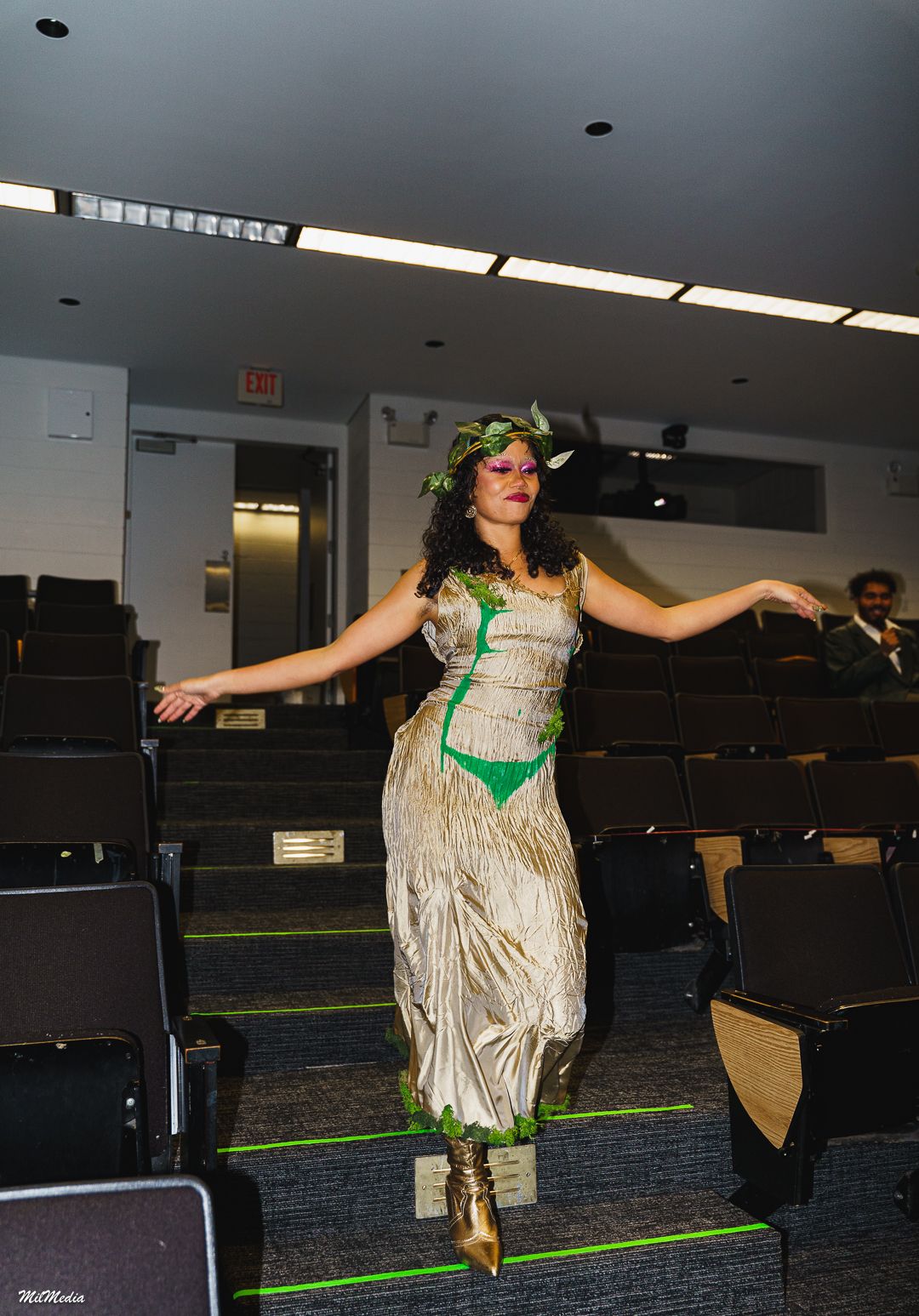 Model in nature-themed gold draped dress and green laurel wreath walks through auditorium.