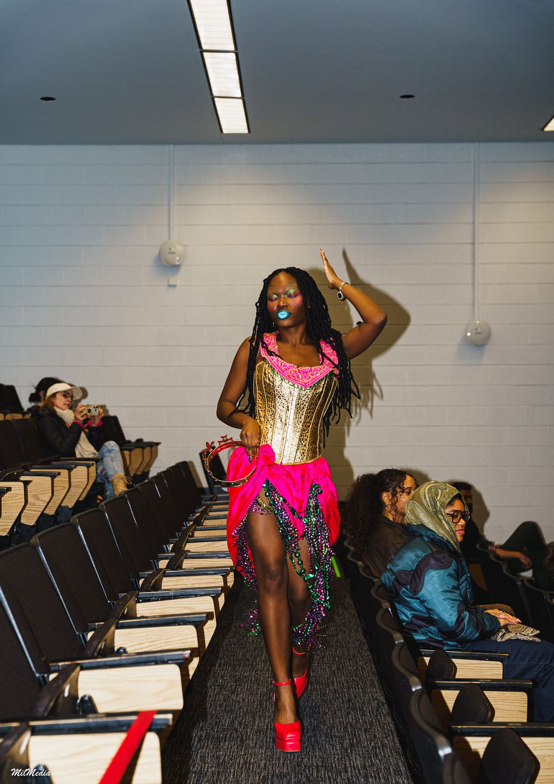 Woman in gold corset and hot pink fringed skirt with red heels walks auditorium runway.