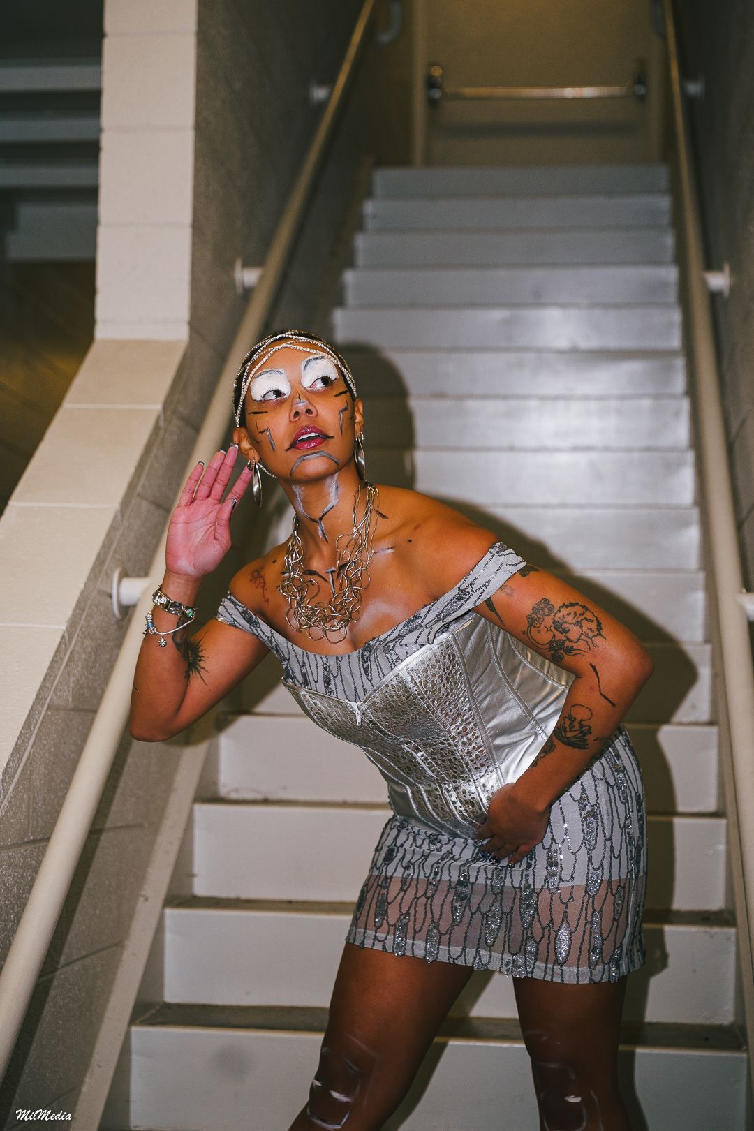A tattooed woman in a silver sequin mini dress and mirrored sunglasses poses on a staircase.