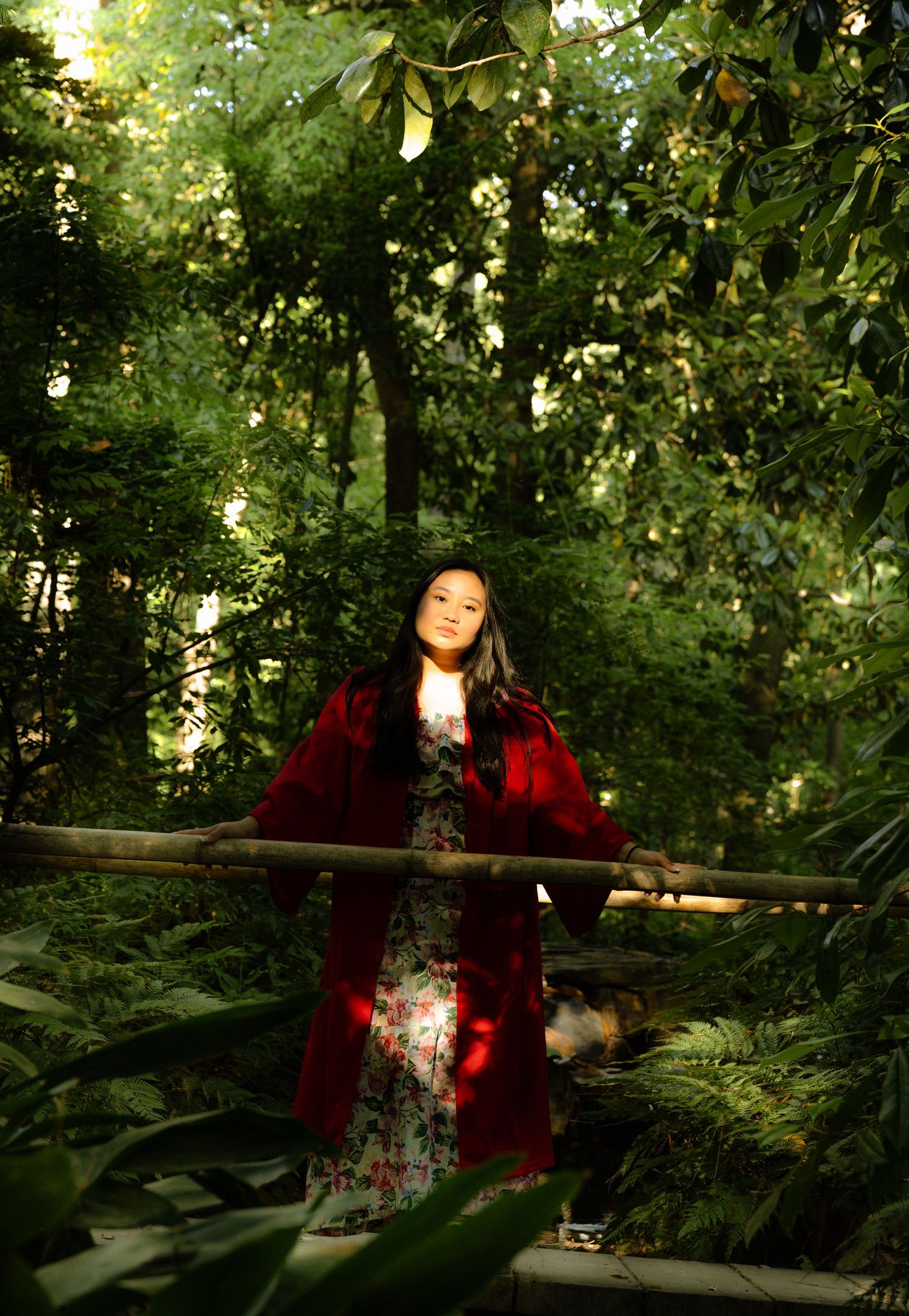 Young woman in red coat and floral dress stands on bamboo bridge in a lush green forest with dappled sunlight.