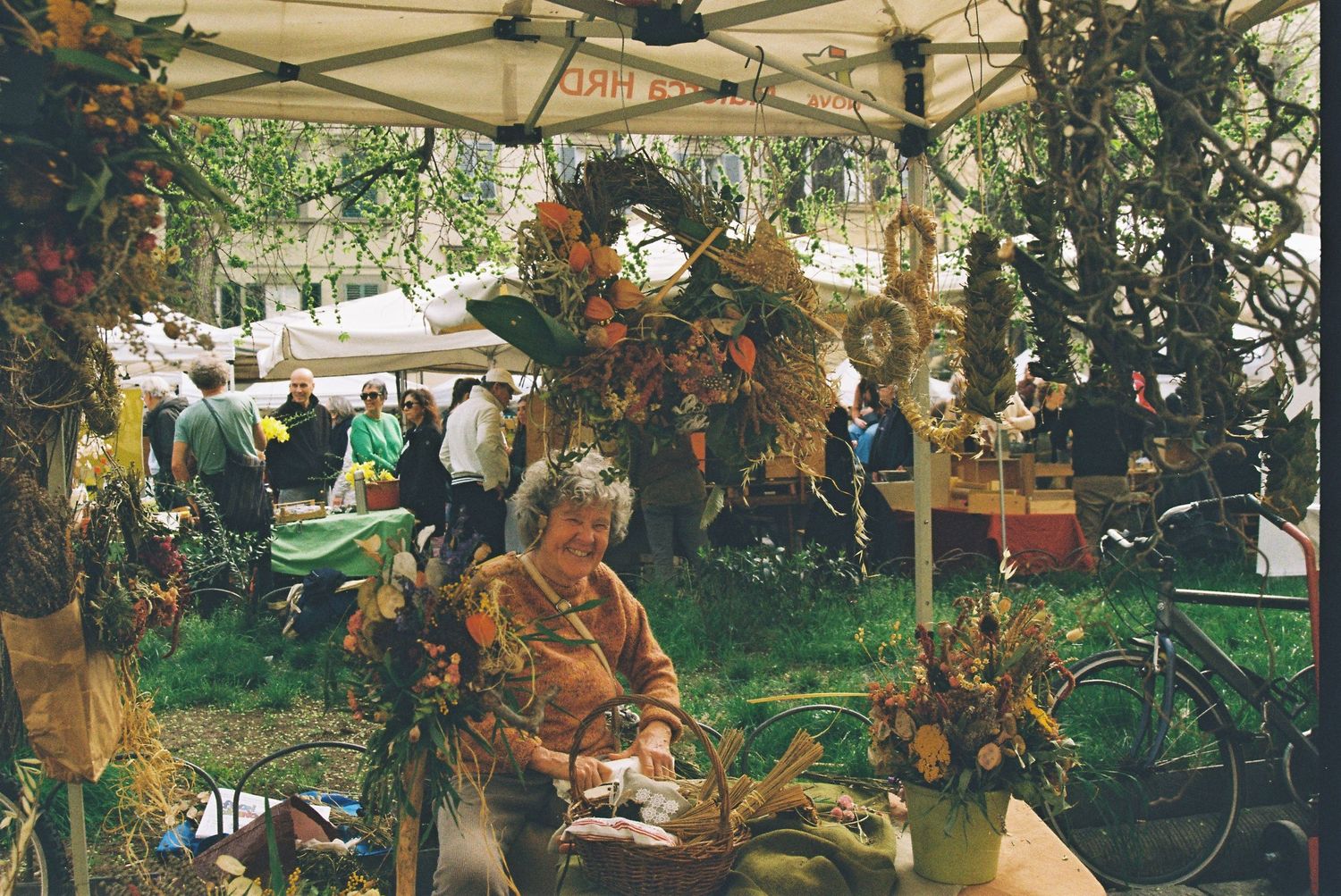 An elderly woman sits at a flower-decorated outdoor market stall surrounded by autumn floral arrangements.