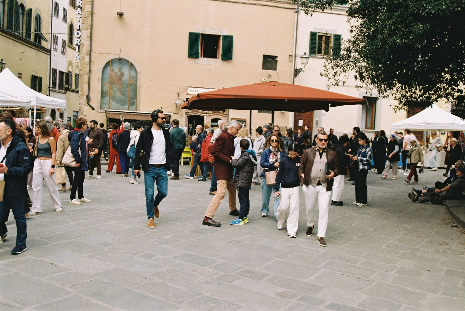 Crowds of people stroll through a busy outdoor market in a sunny Italian piazza with white vendor tents.