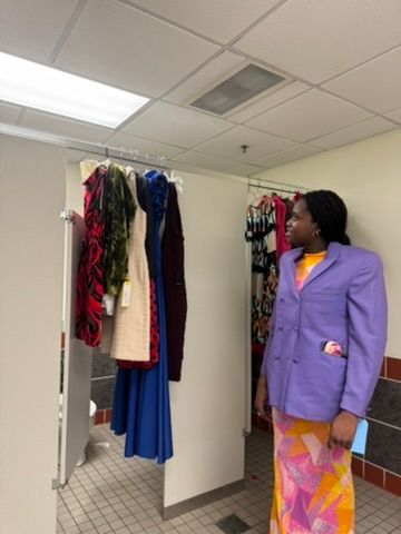 Woman in purple blazer and colorful skirt reviews clothing rack with vibrant garments backstage at fashion event.