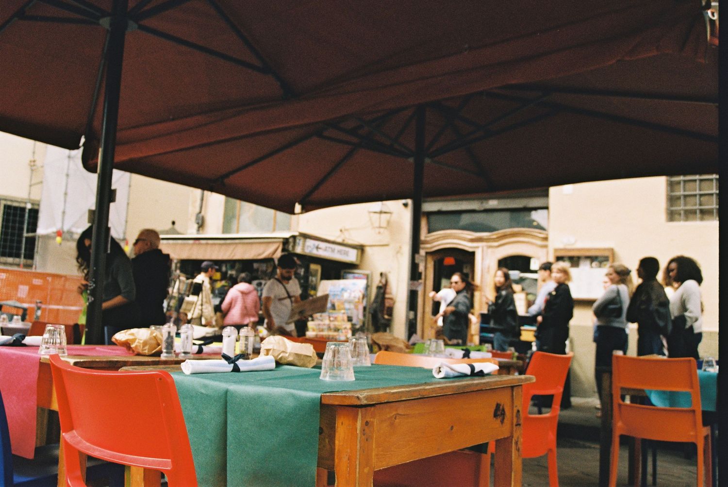 Outdoor market scene with colorful chairs, tables under umbrellas, and people browsing vendor stalls.