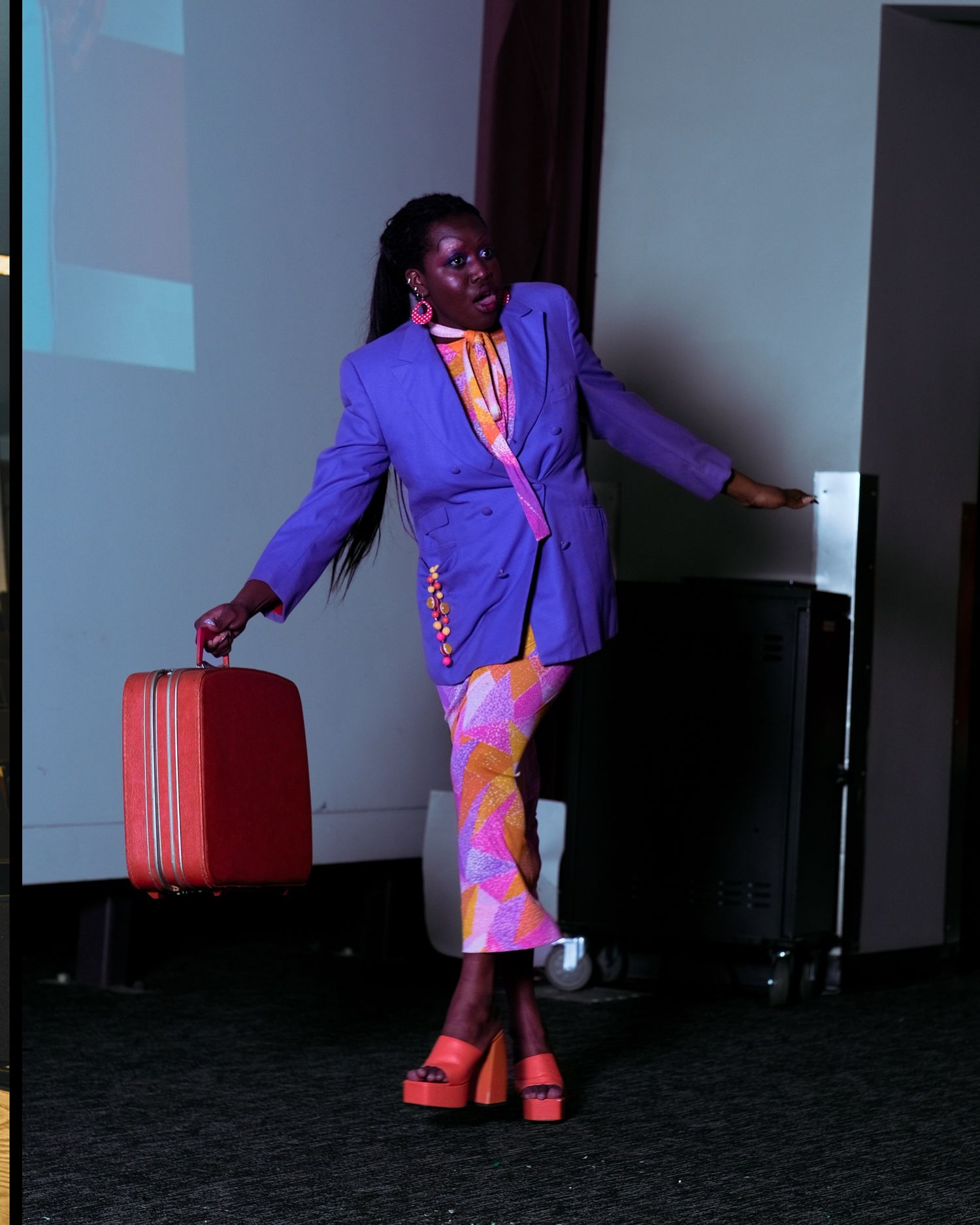 A model walks a runway in a purple blazer, colorful pants, and red platform heels carrying a red suitcase.