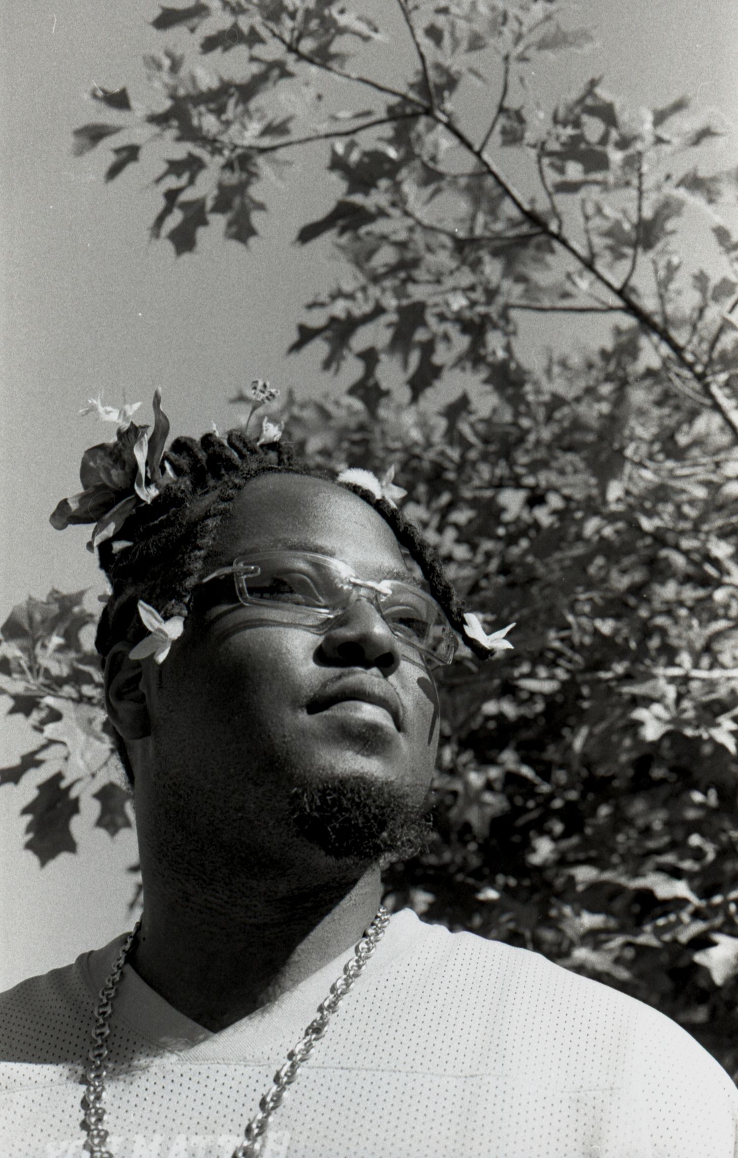 Black and white portrait of a Black man with flowers in his hair and glasses looking upward outdoors.
