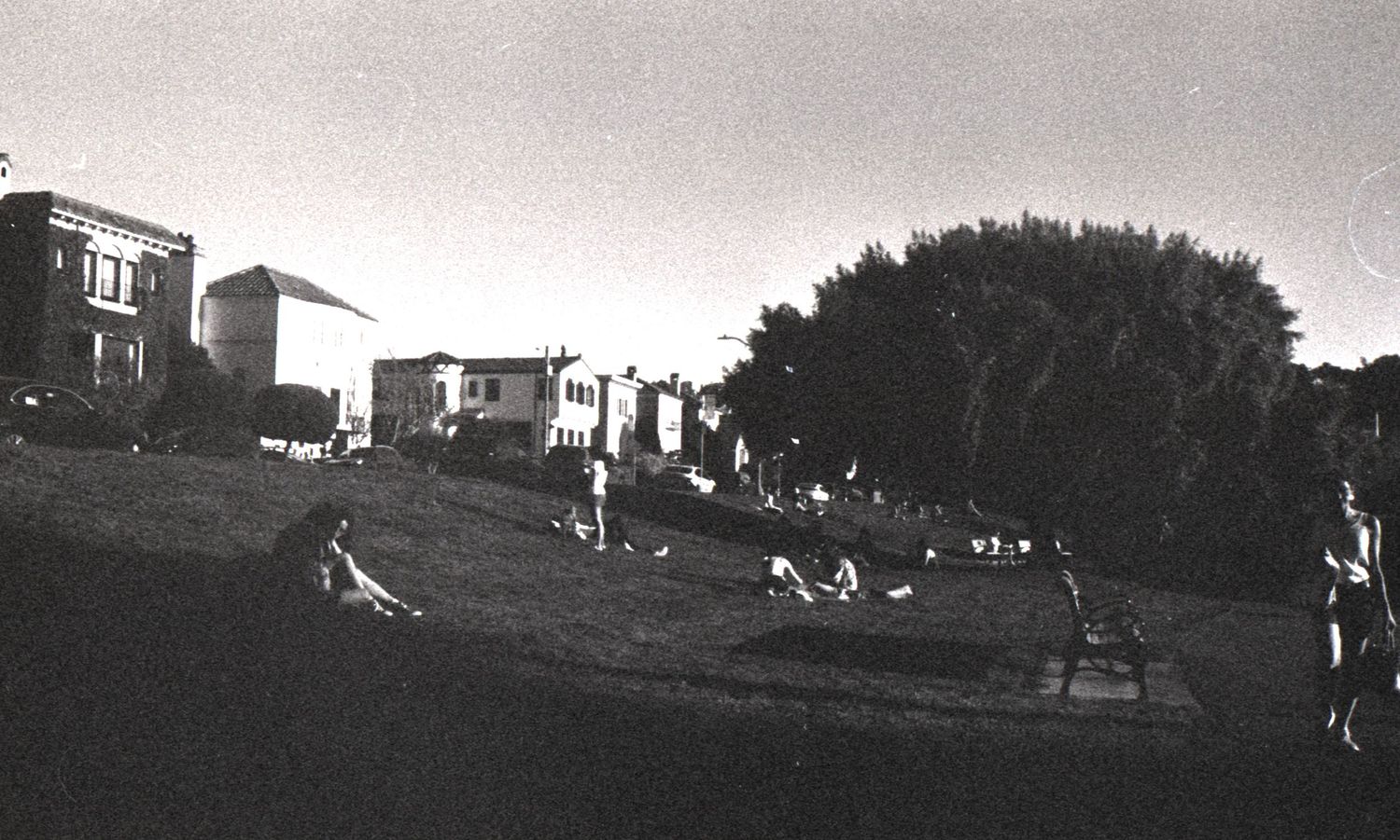 Black and white panoramic photo of people relaxing on a grassy hill with urban buildings behind.