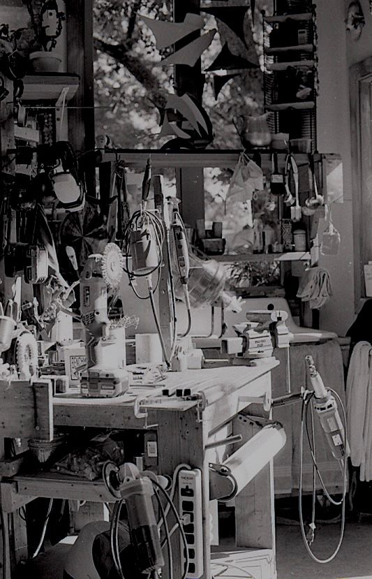 Black and white image of a cluttered artist workshop workbench with tools and equipment.