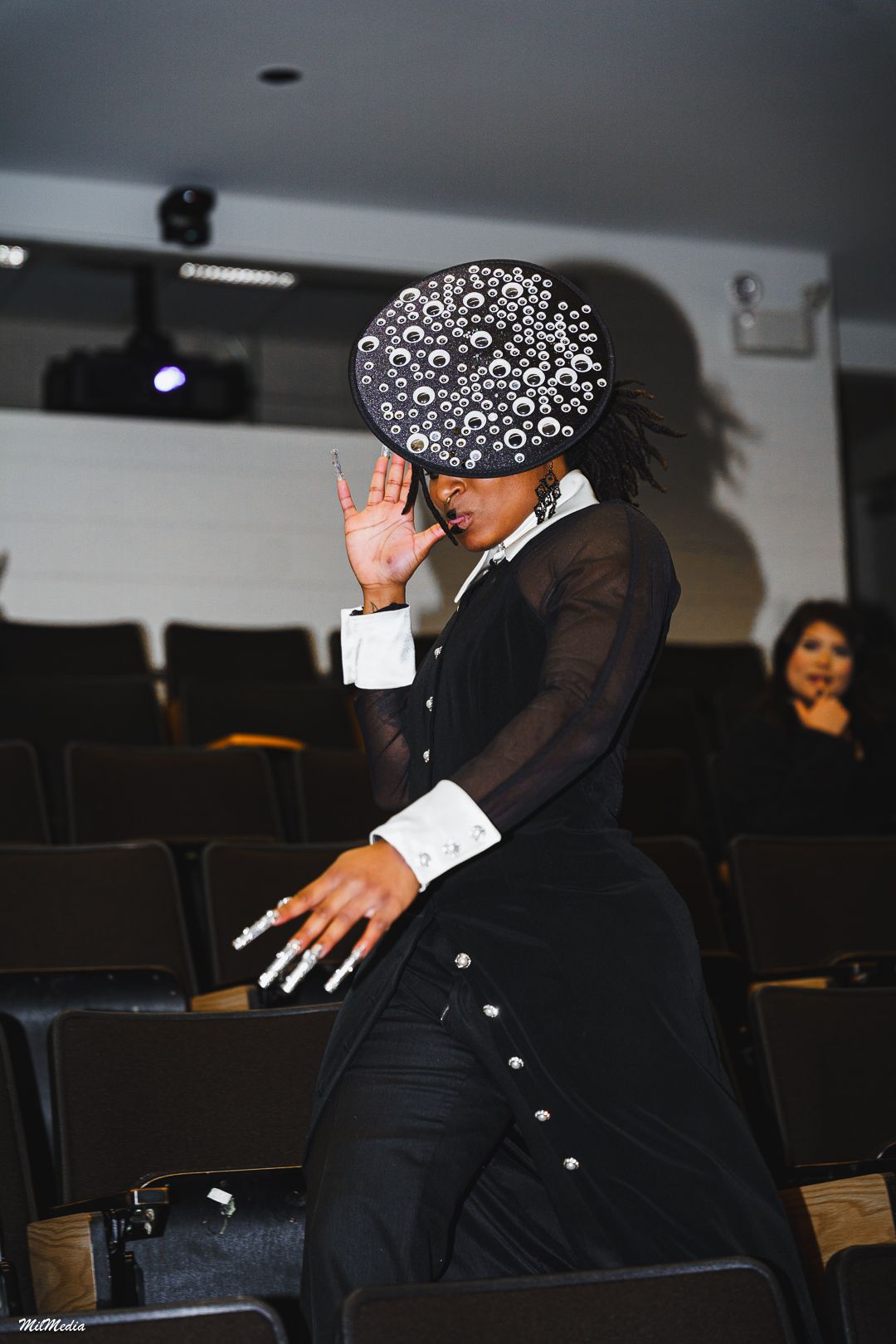 A model in a sheer black gown and jeweled hat poses dramatically in an auditorium.
