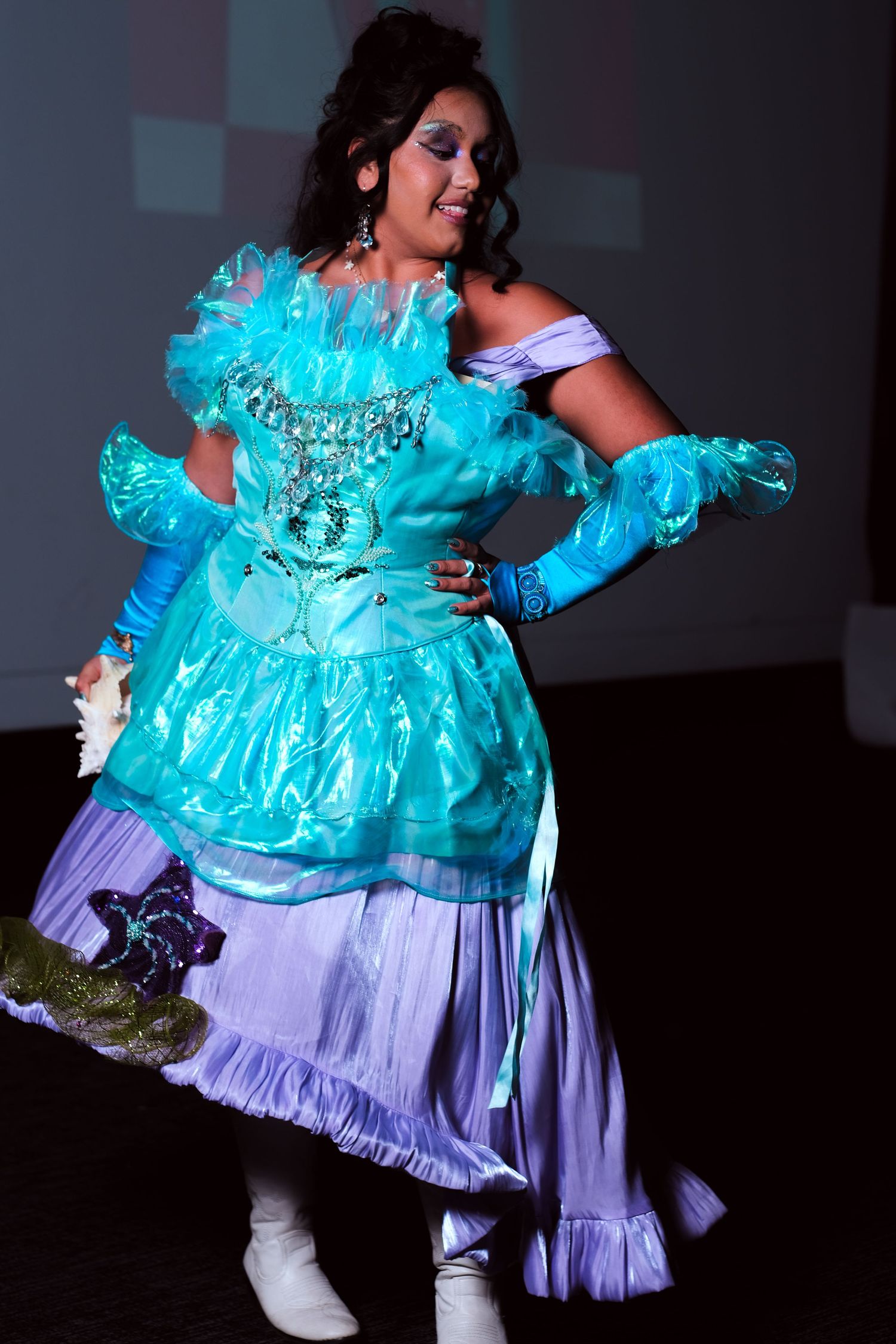 Woman in elaborate turquoise and lavender ball gown with corset performs on a dimly lit stage.