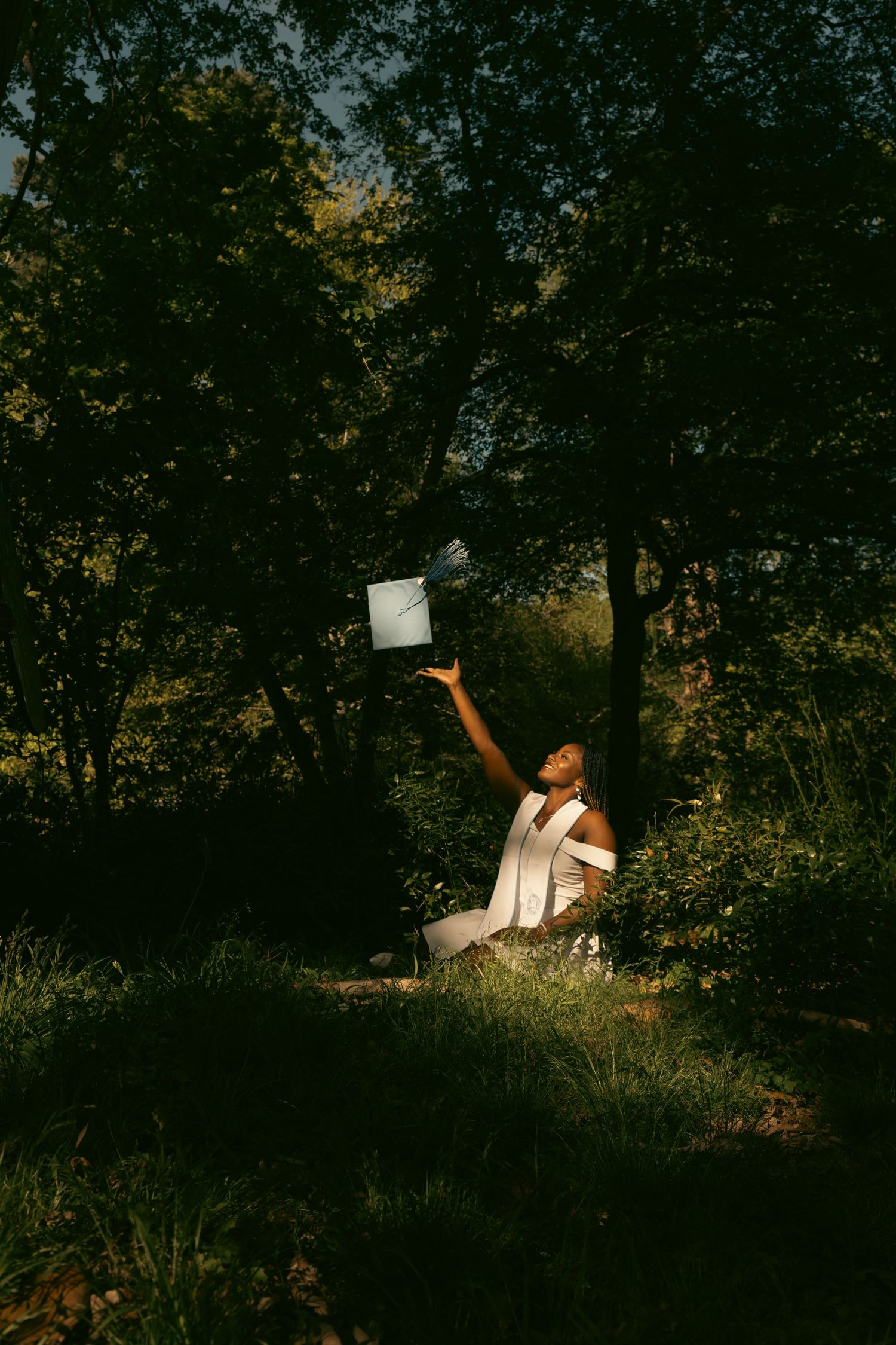 Woman in white dress tosses graduation cap in sunlit forest clearing surrounded by green trees.