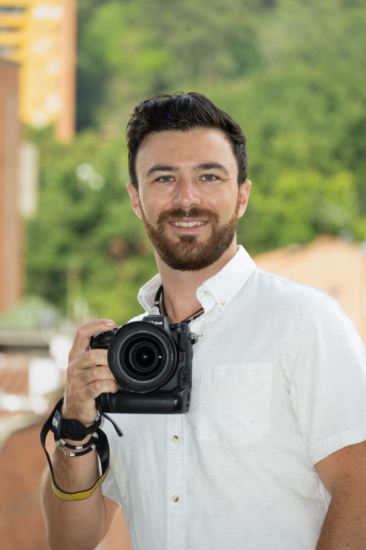 Professional photographer holding DSLR camera while wearing white shirt against blurred outdoor background.