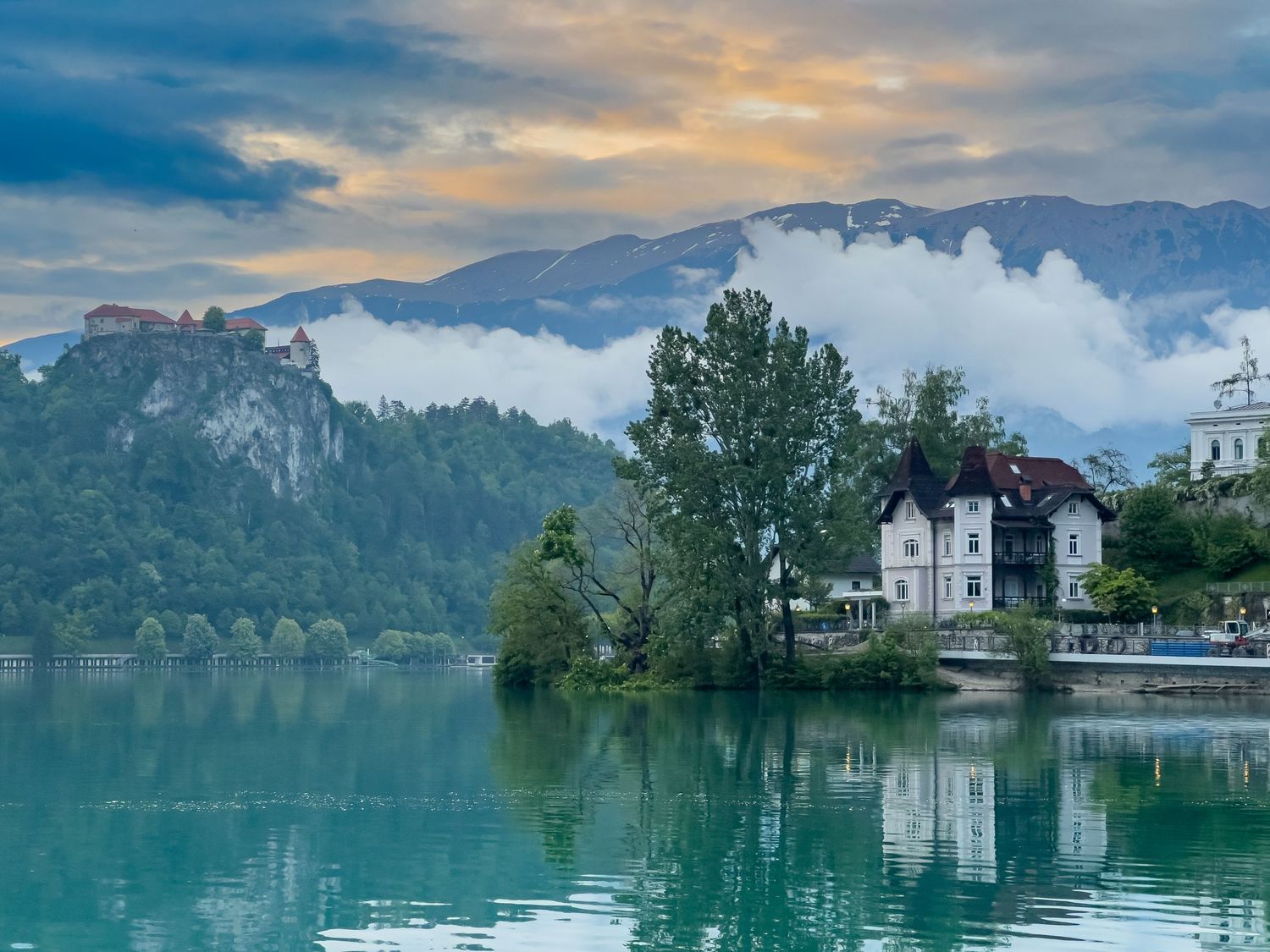 Lake Bled in Slovenia reflects a dramatic sunset sky with mountains, trees, and buildings along its tranquil shoreline.