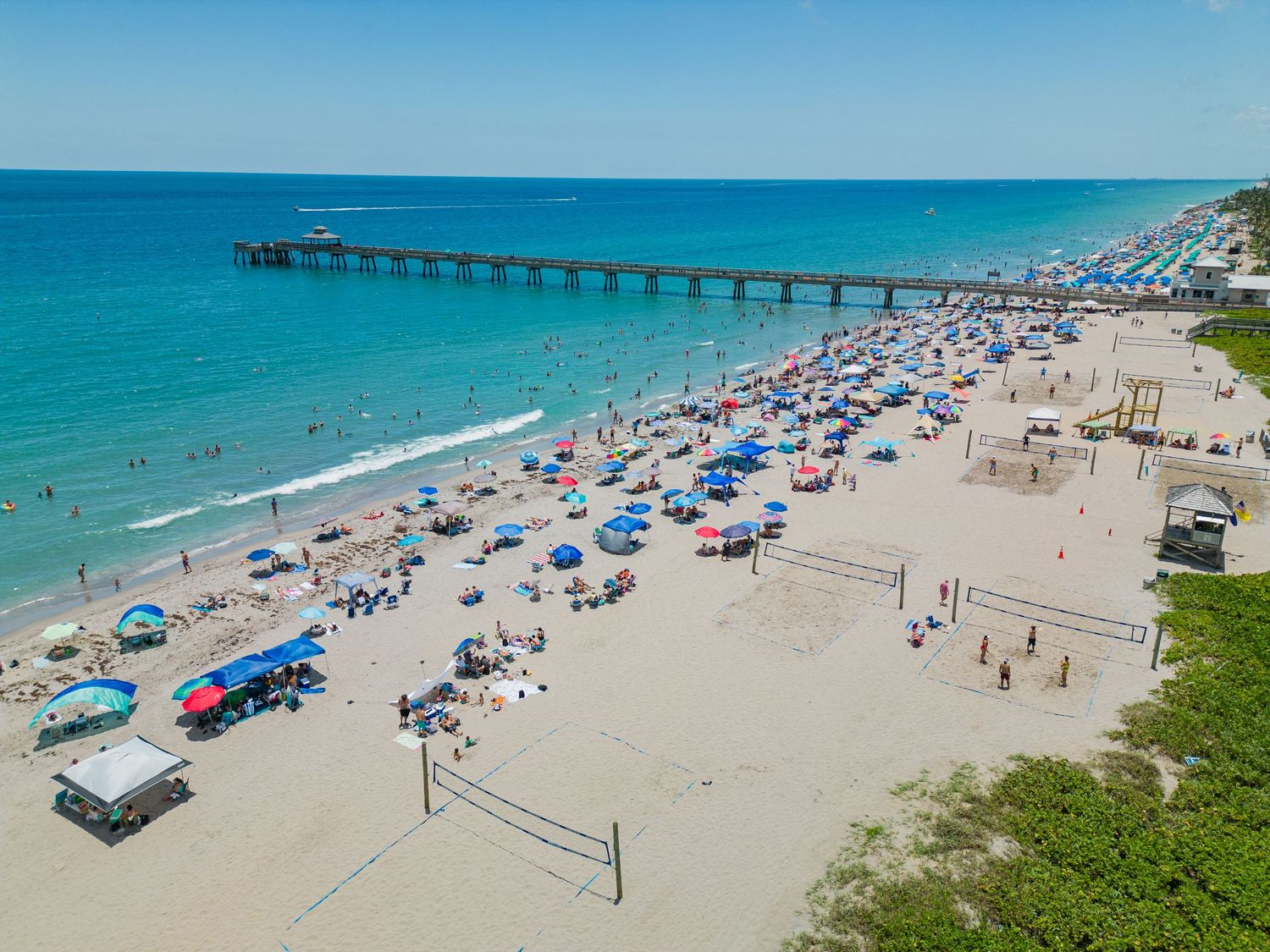 Aerial view of Deerfield Beach pier in Florida with turquoise waters, sandy shores and colorful beach umbrellas.