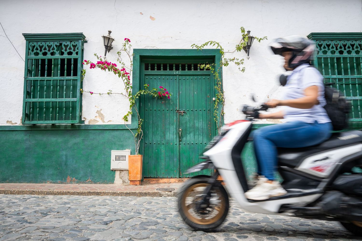 A series of green shuttered windows and doors line a white wall as scooters pass by on a cobblestone street.