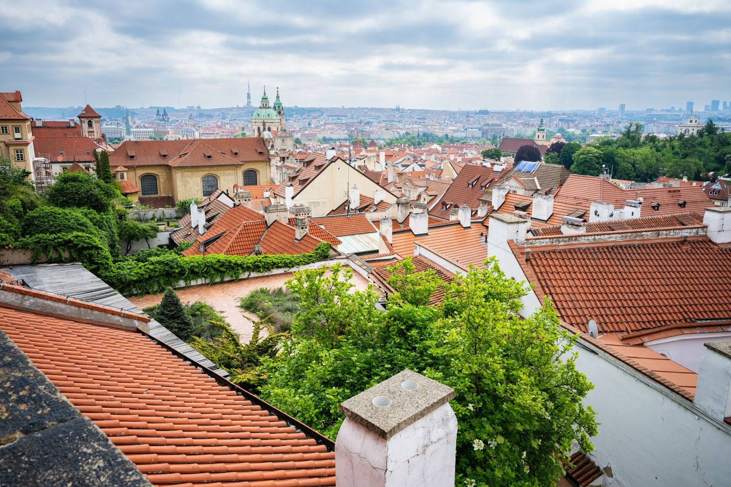 Panoramic view of Prague's historic skyline with orange terracotta rooftops and church spires against a cloudy blue sky.