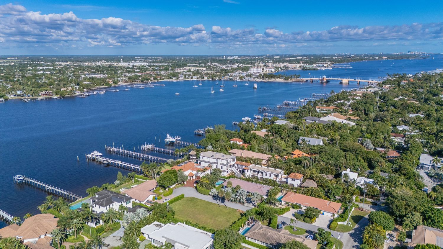 Aerial panoramic view of coastal waterfront with boats, docks, and residential buildings under bright blue sky.