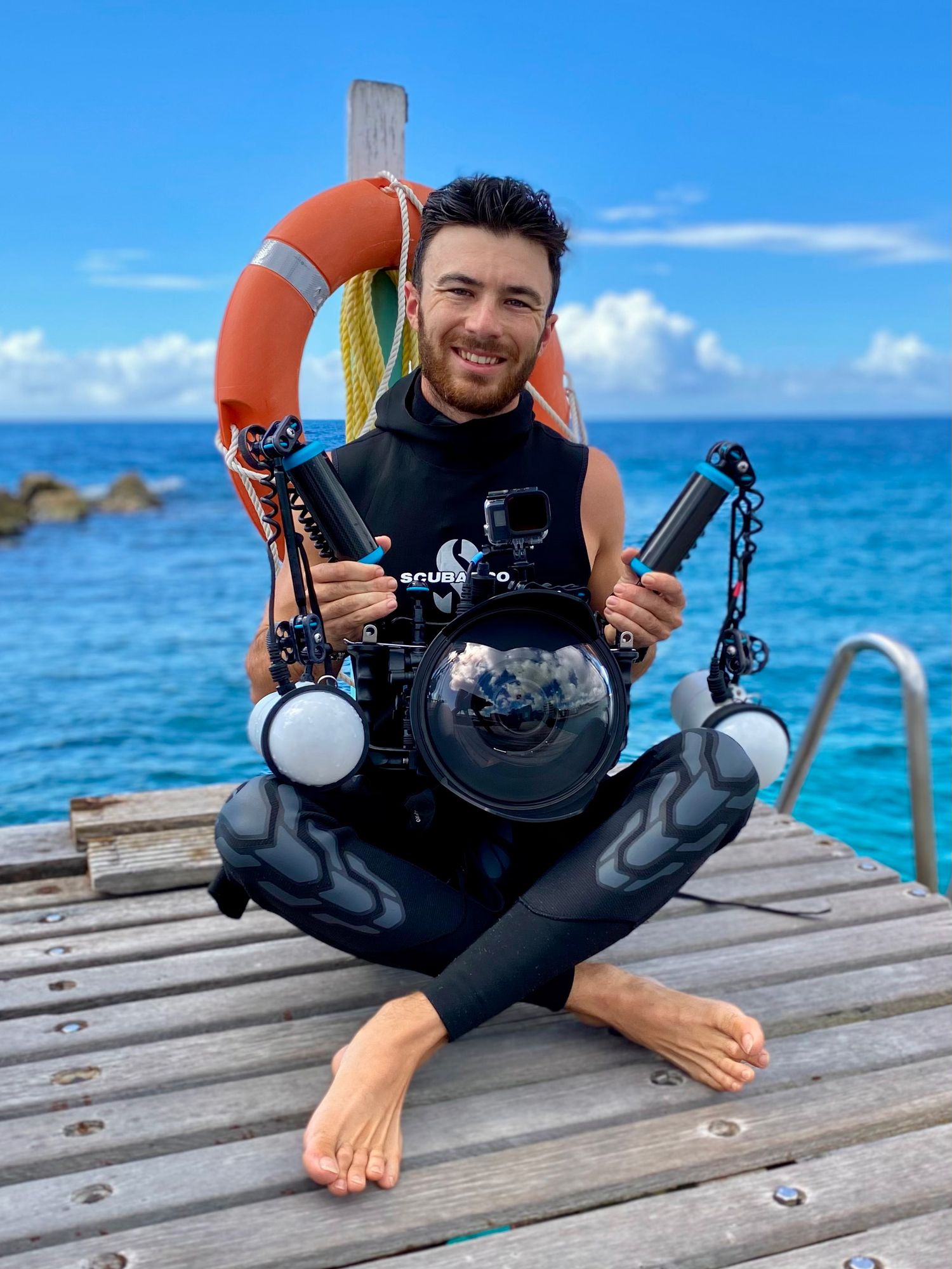 Scuba diver with underwater photography equipment resting on wooden dock over tropical blue waters.