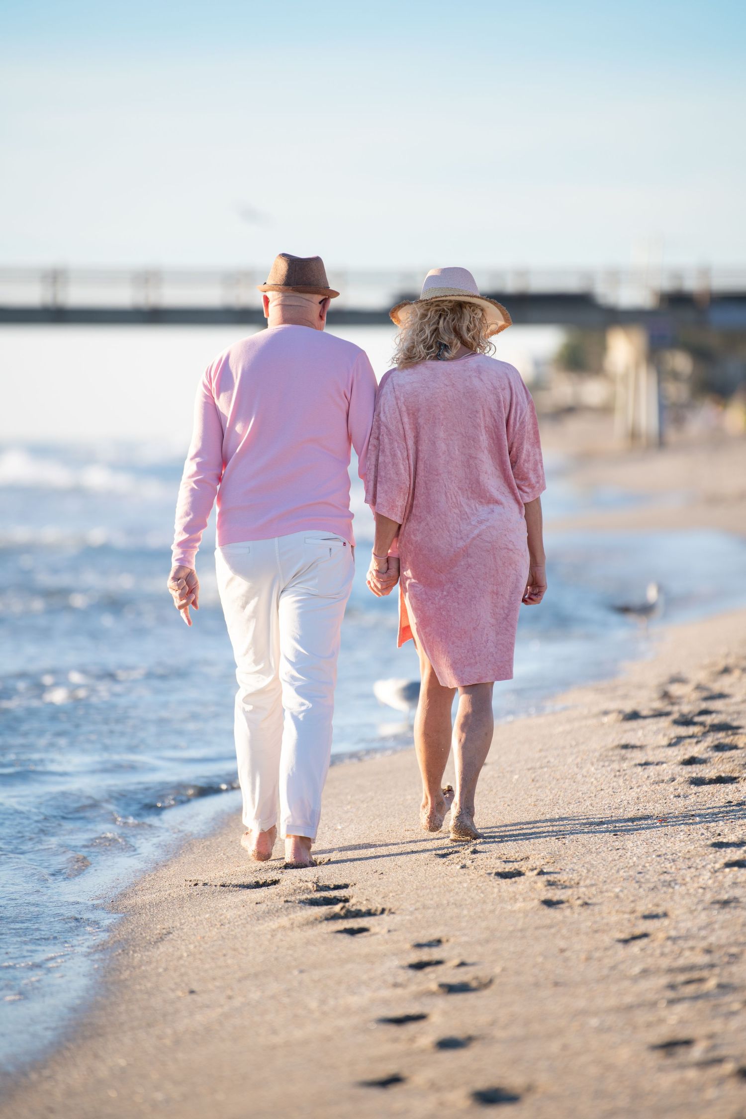 Couple in coordinated pink and white outfits walking together along a sunny beach shoreline.