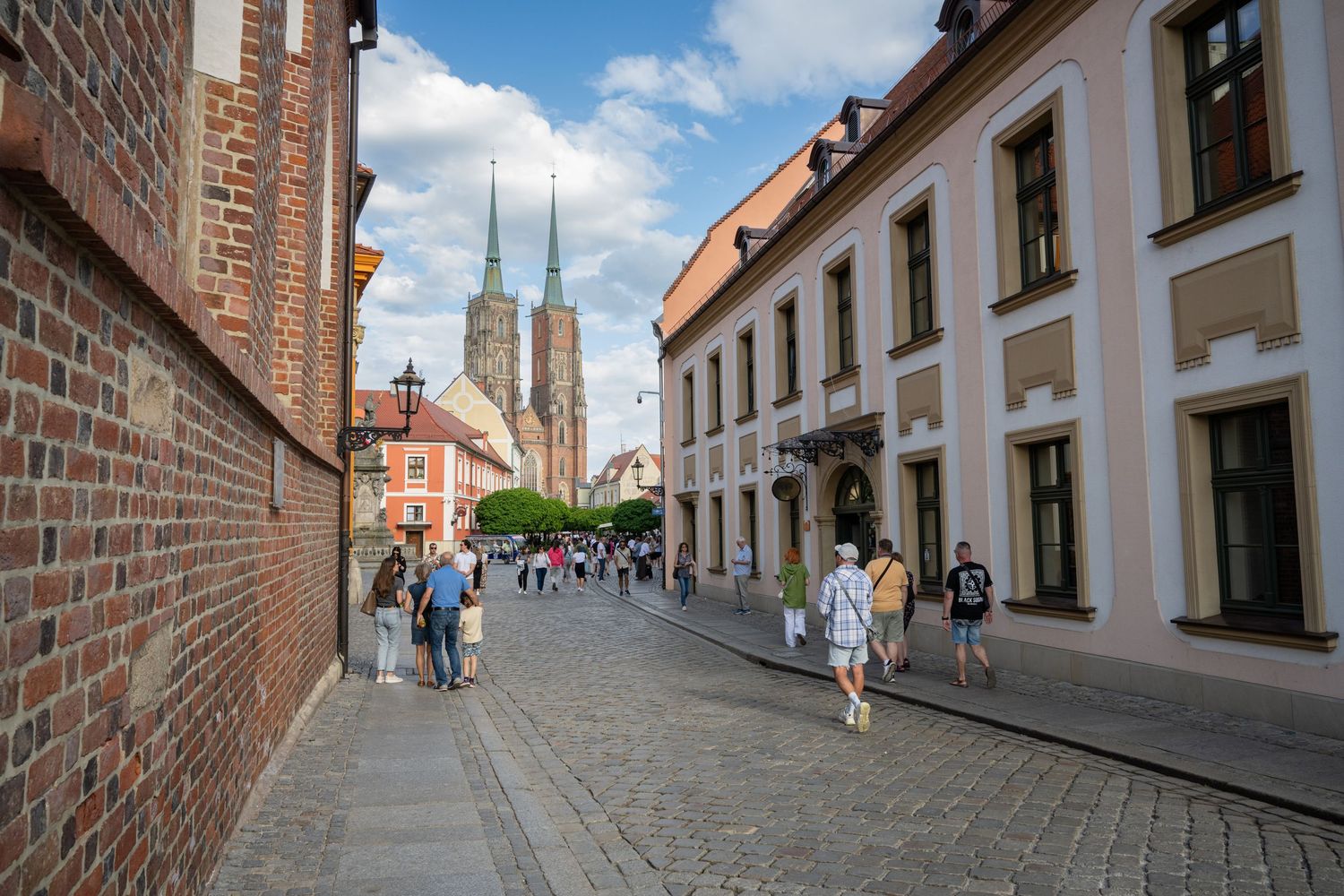 Narrow cobblestone street lined with historic brick buildings and spires visible in the distance on a sunny day.