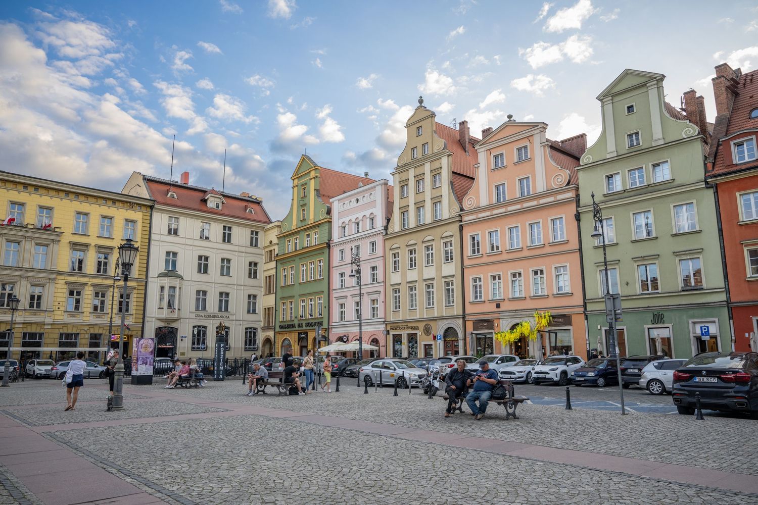 Colorful historic buildings line the town square in Opole, Poland with traditional European architecture and parked cars.