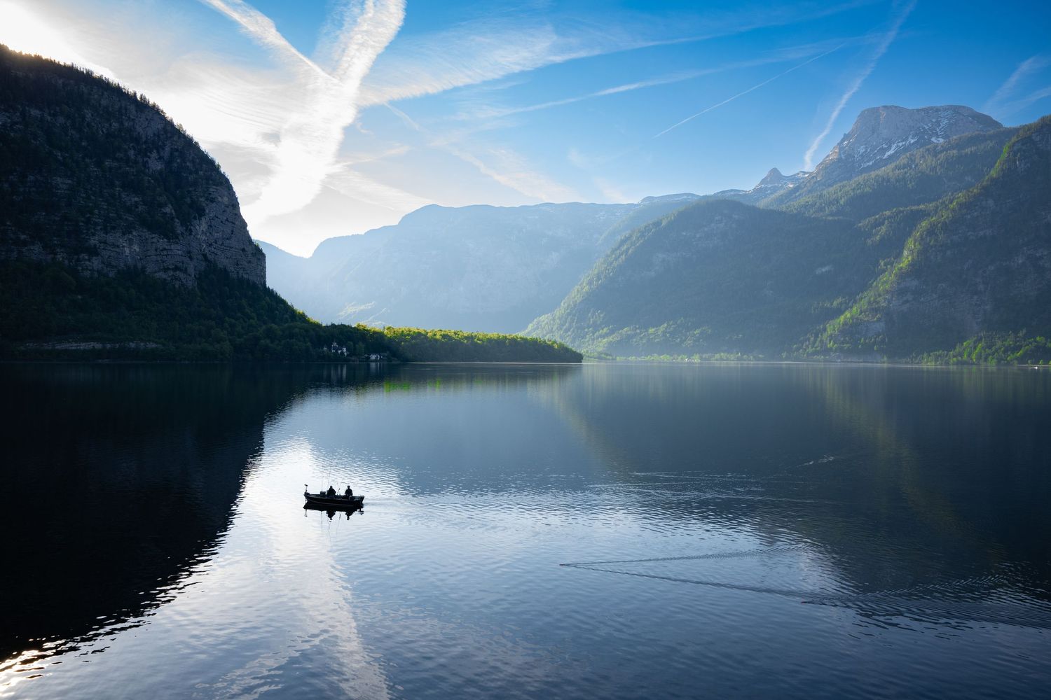 Serene mountain lake surrounded by steep cliffs reflects clouds and sunlight on calm misty waters at dawn.