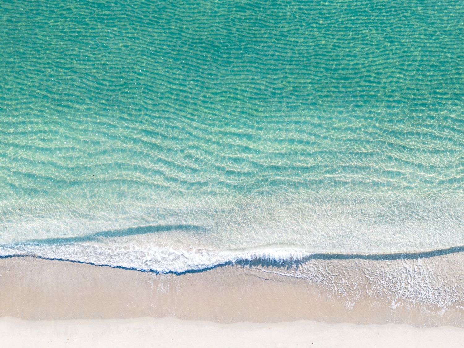 Aerial view of turquoise ocean waves gently rolling onto a pristine sandy beach with white foam patterns.