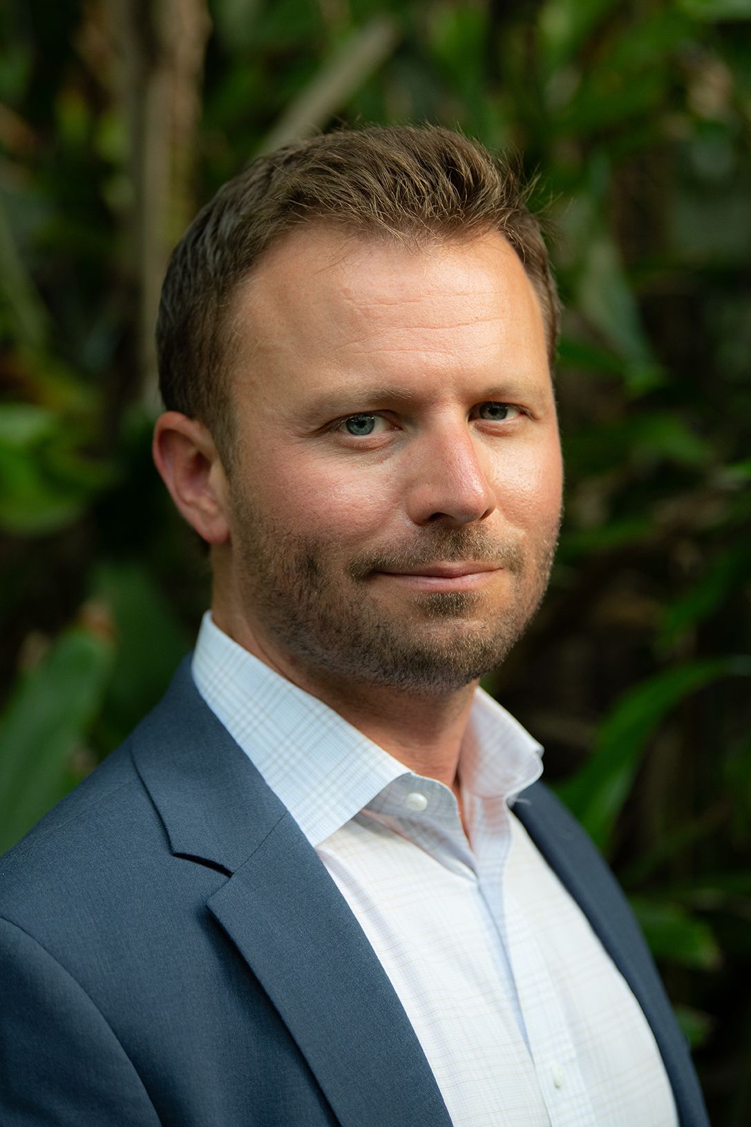 Professional headshot of person in navy suit jacket and light blue collared shirt against green foliage background.