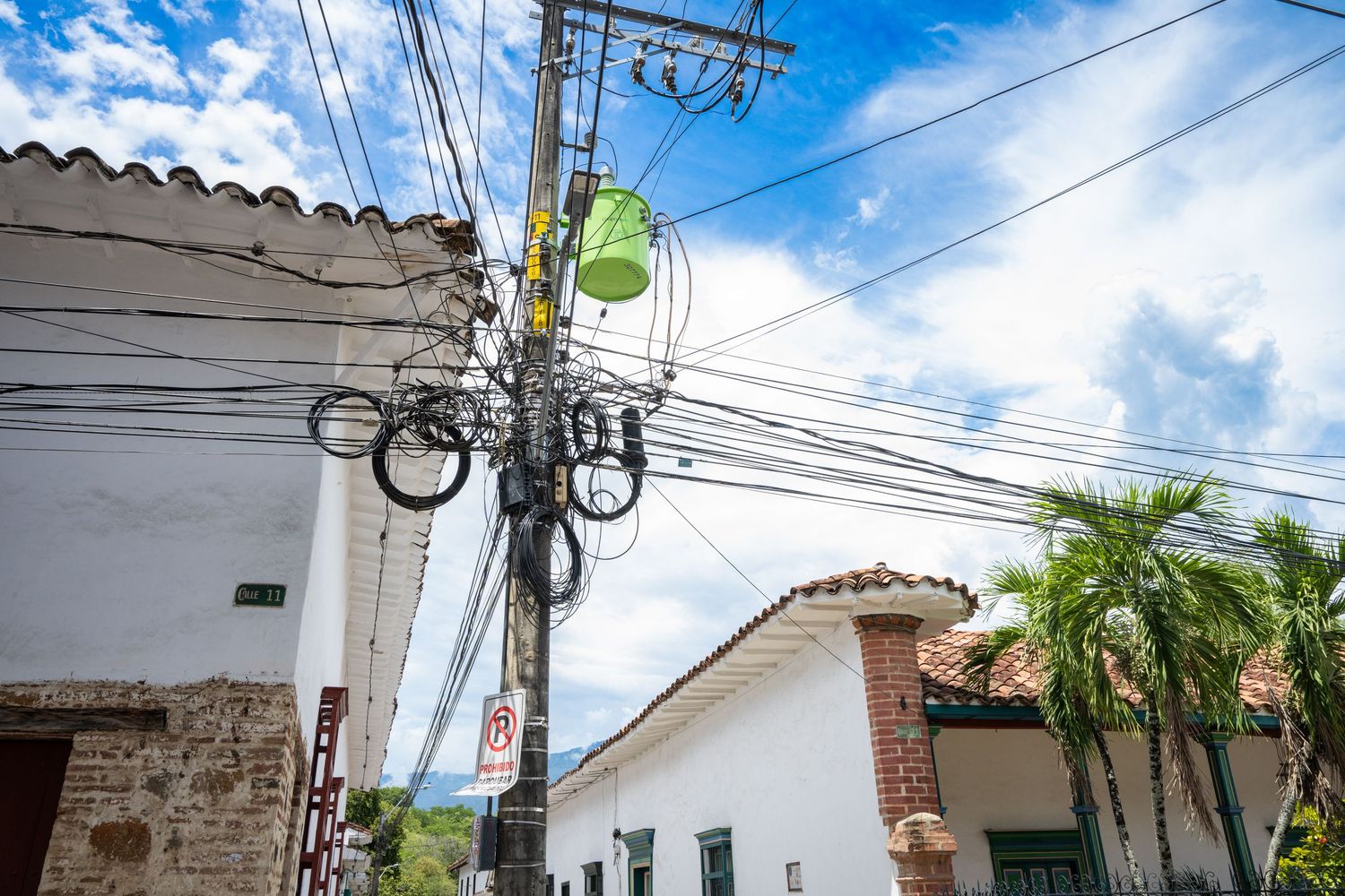 Tangled power lines and transformers against blue sky with white buildings and palm trees in tropical setting.