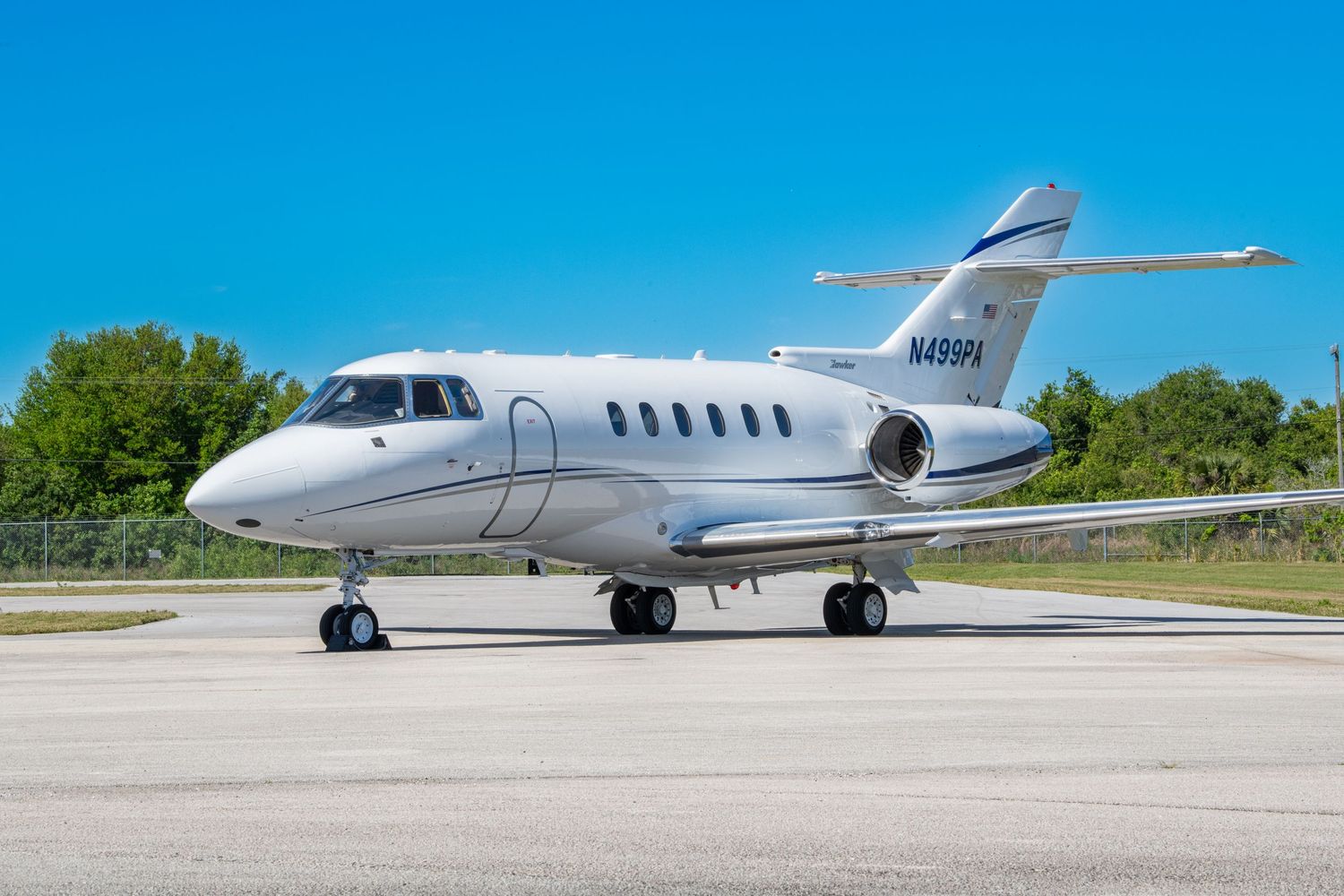 A sleek white Hawker 800 business jet parked on a concrete tarmac with green trees in the background.